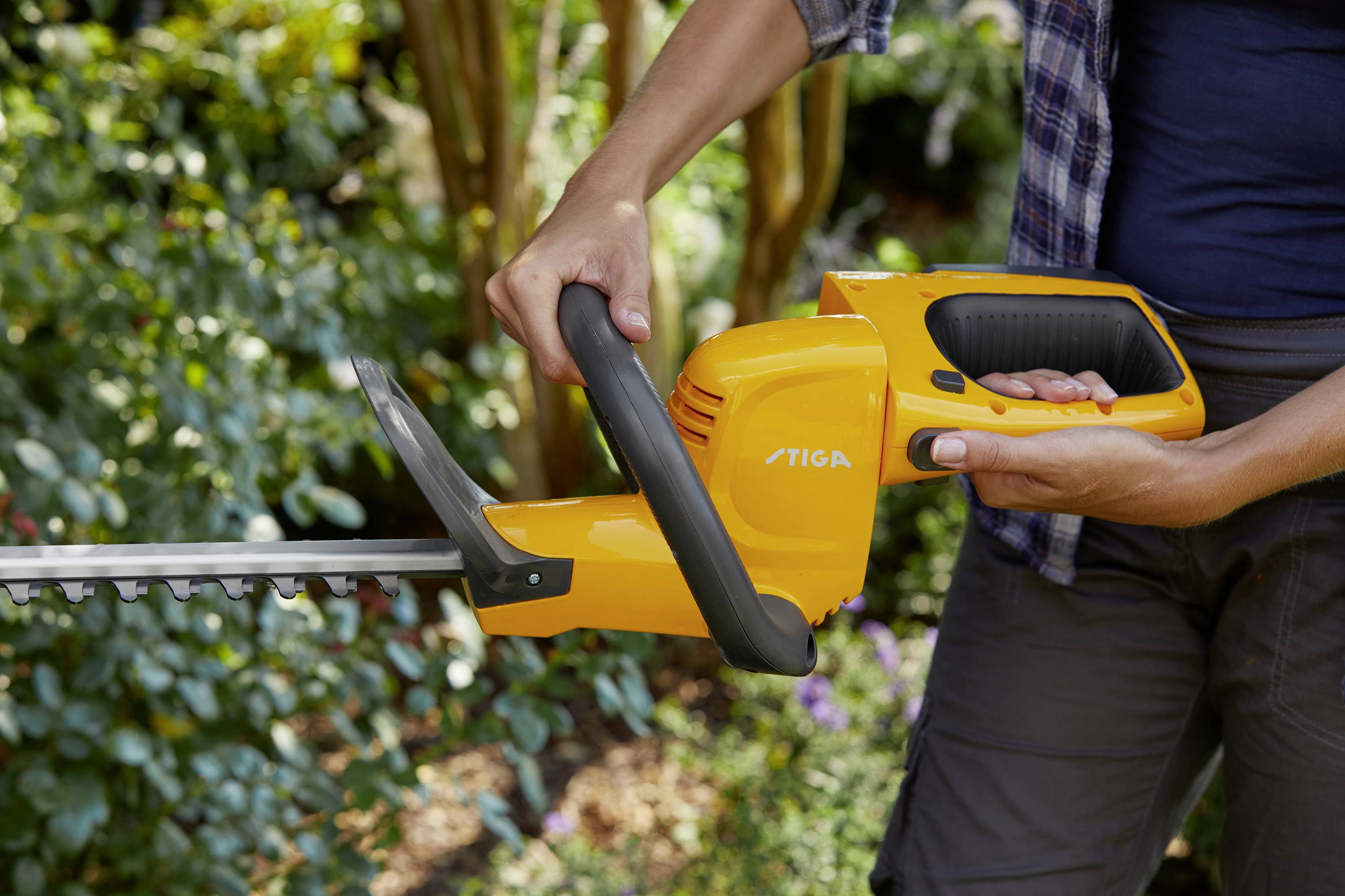 A person is operating a bright yellow electric hedge trimmer, surrounded by green plants in the garden.