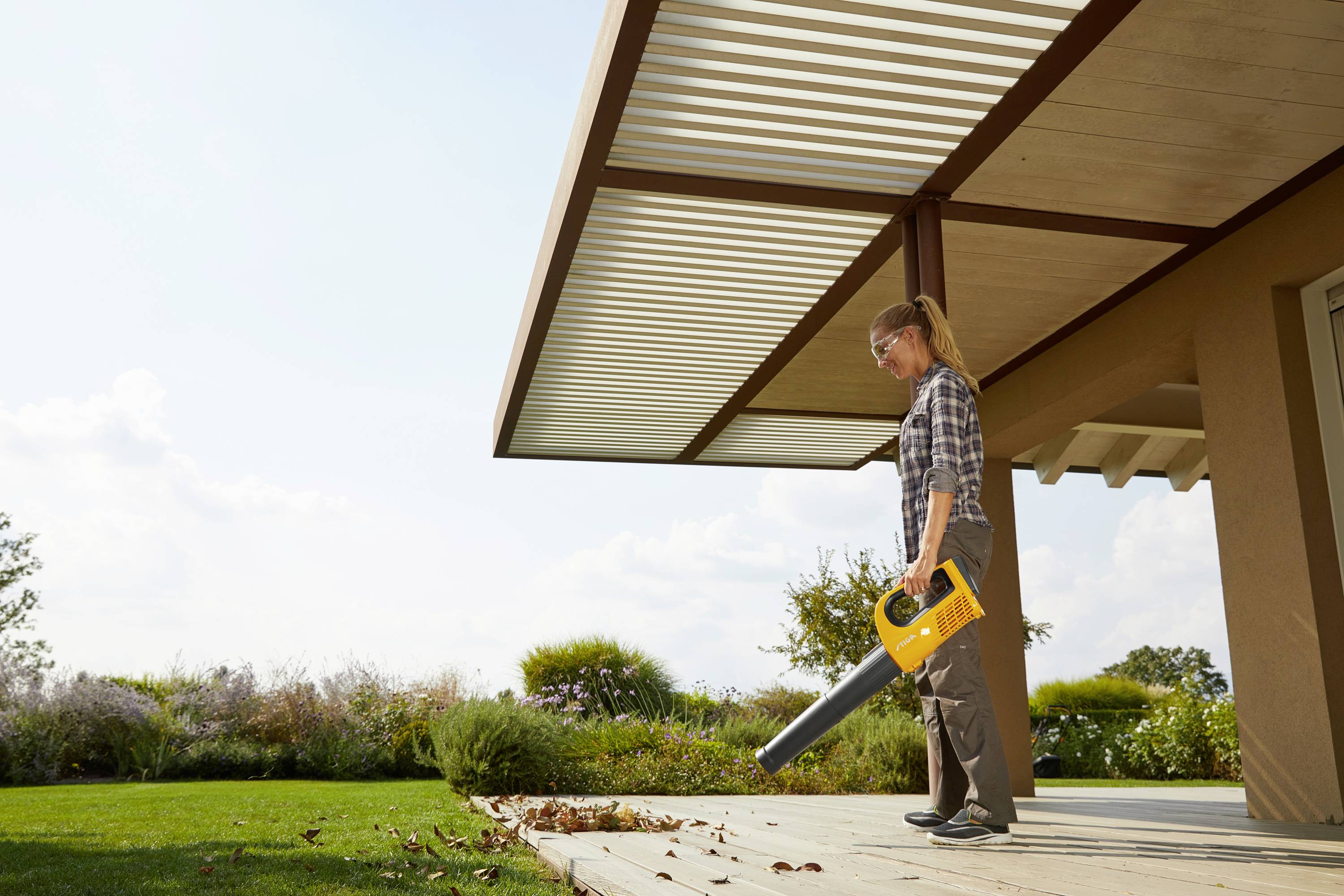 A woman is using a yellow leaf blower to remove leaves from a patio. Trees and bushes can be seen in the background.