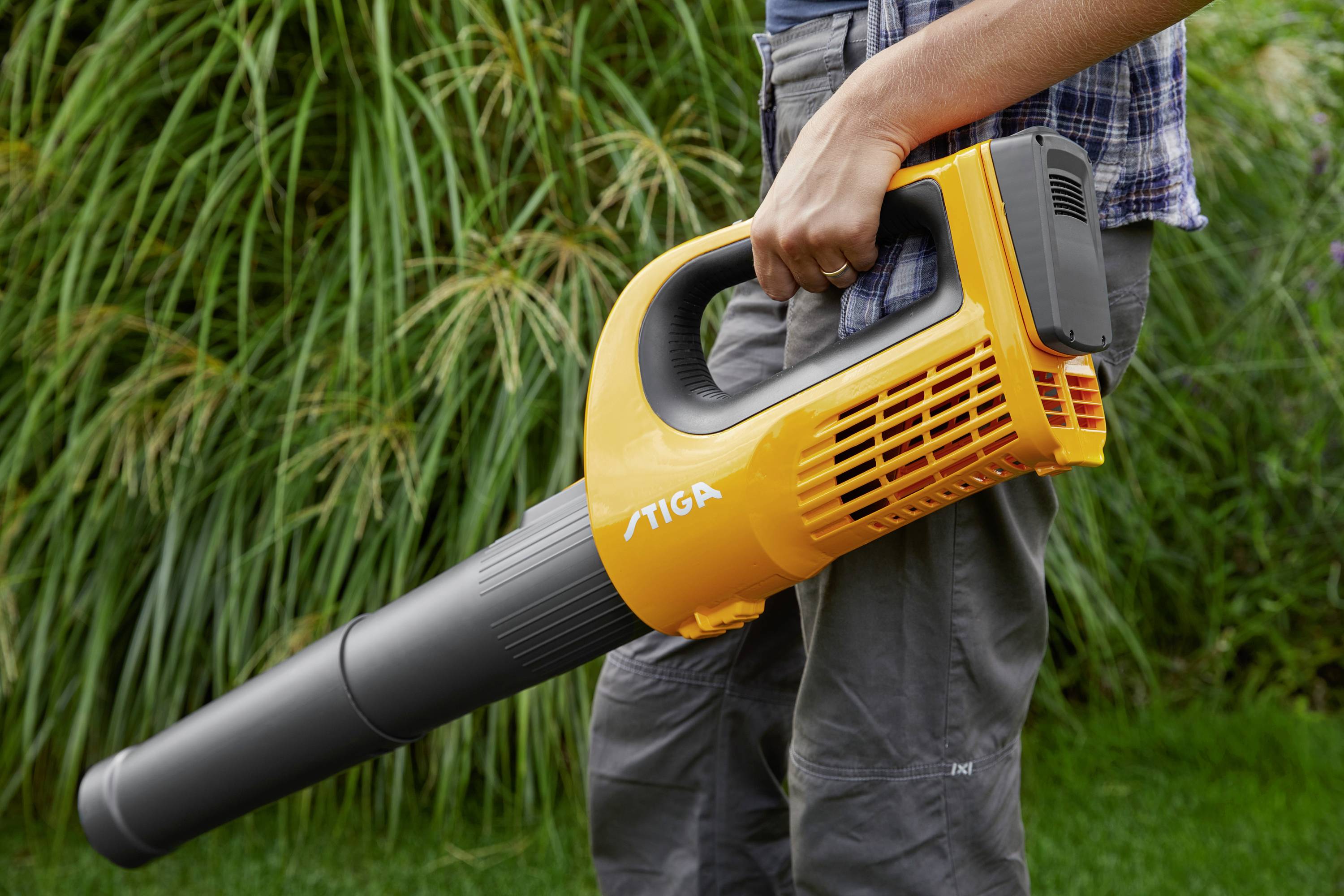 A person is holding a yellow leaf blower while standing in a garden next to tall grasses.