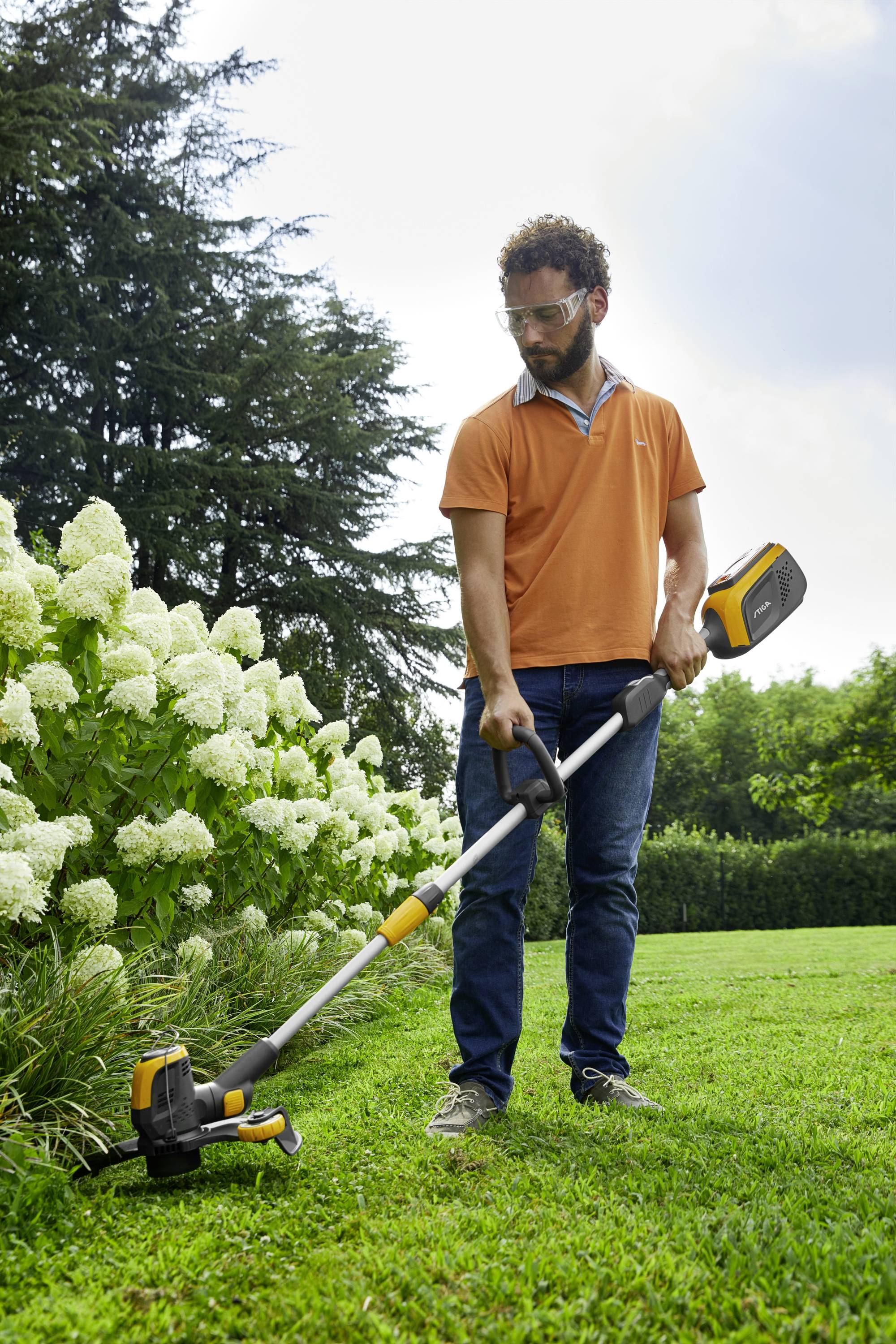 A man in an orange shirt is trimming the grass with an electric lawn trimmer next to white flowers in a garden.