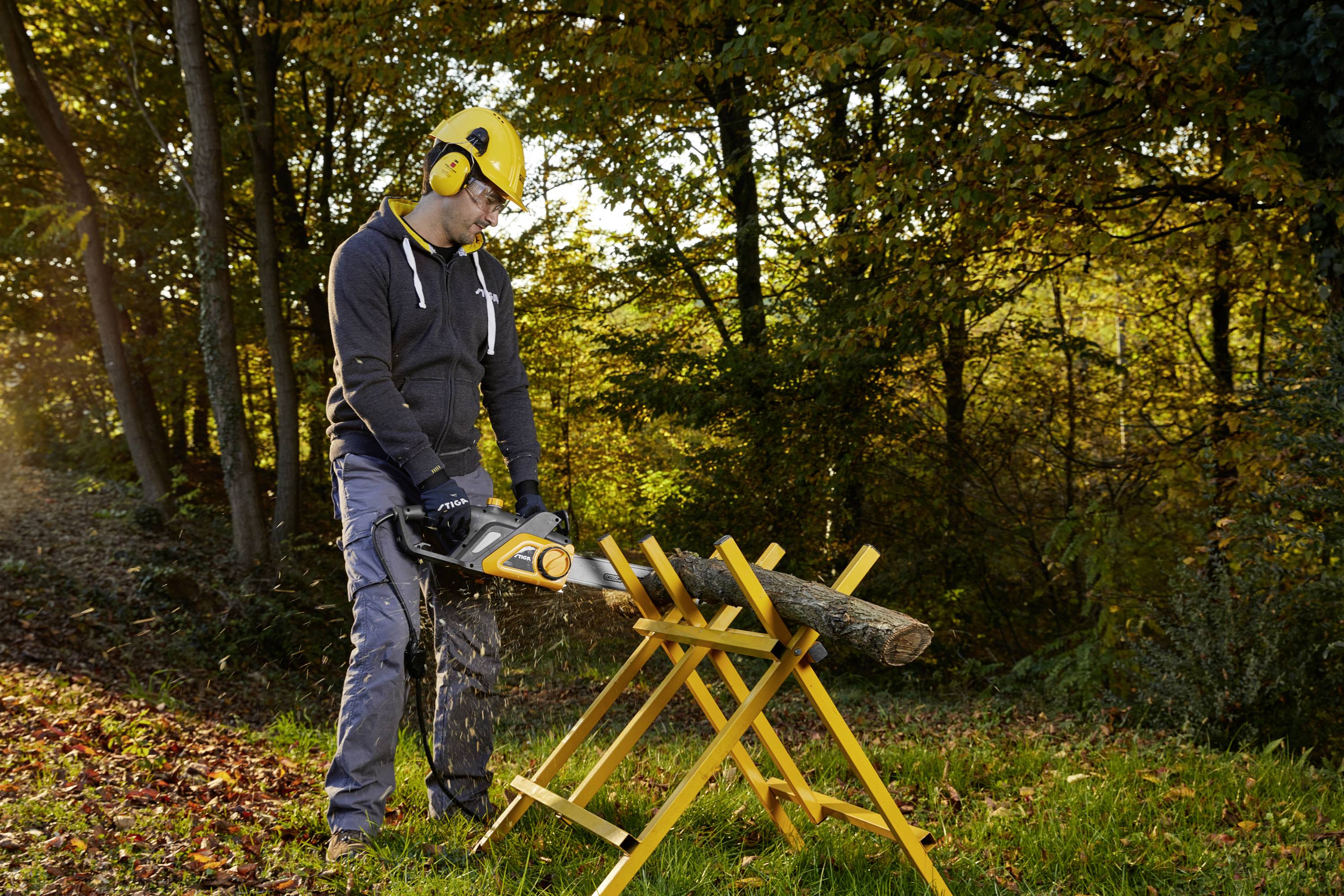 A worker is sawing a tree trunk on a sawbuck with a chainsaw in the forest. He is wearing protective clothing, including a helmet and ear defenders. The surrounding environment reflects an autumnal setting.