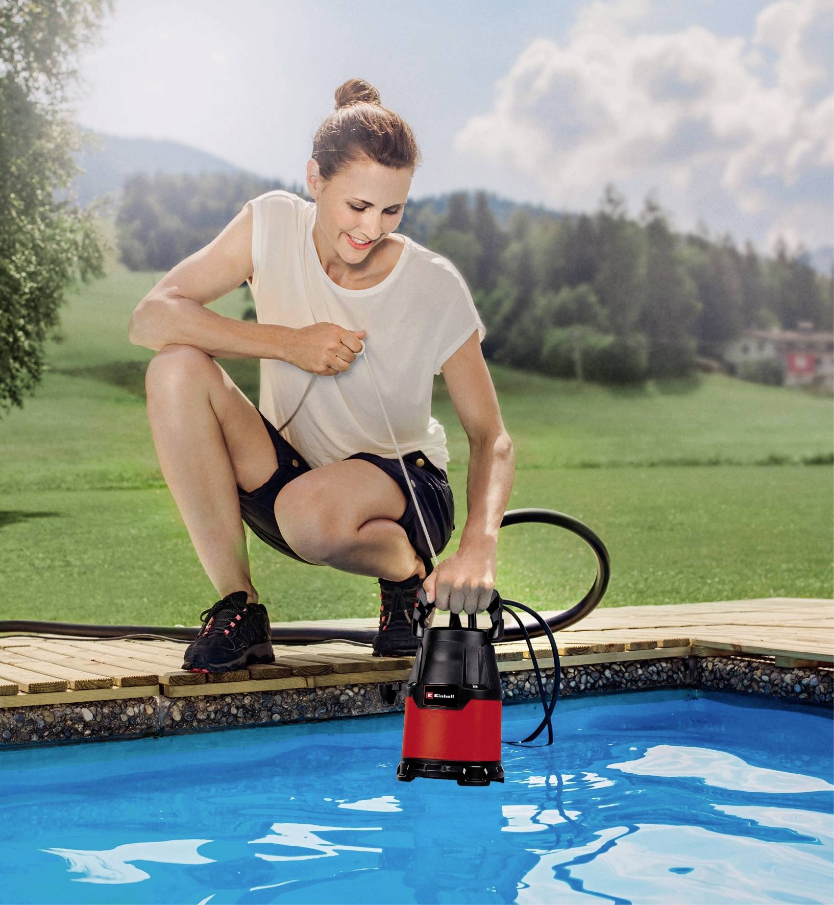 A person is kneeling at the edge of a pool and operating a submersible pump to drain water. A green lawn is visible in the background.