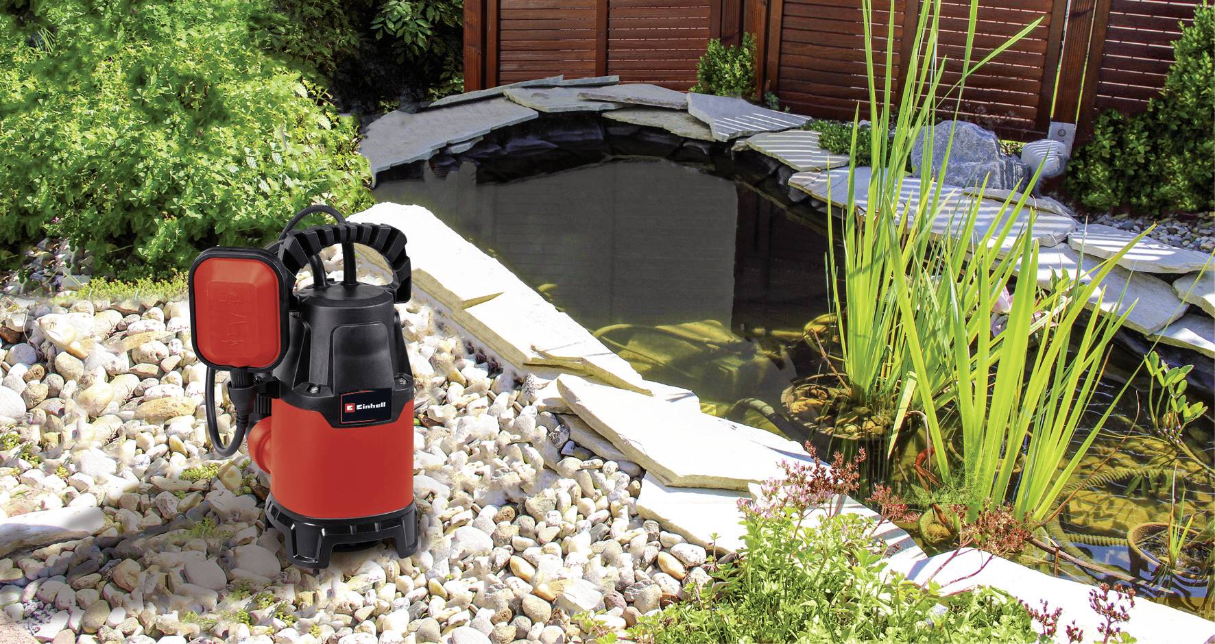 A red water filter or pump device stands beside a garden pond, surrounded by stones and plants, under the open sky.
