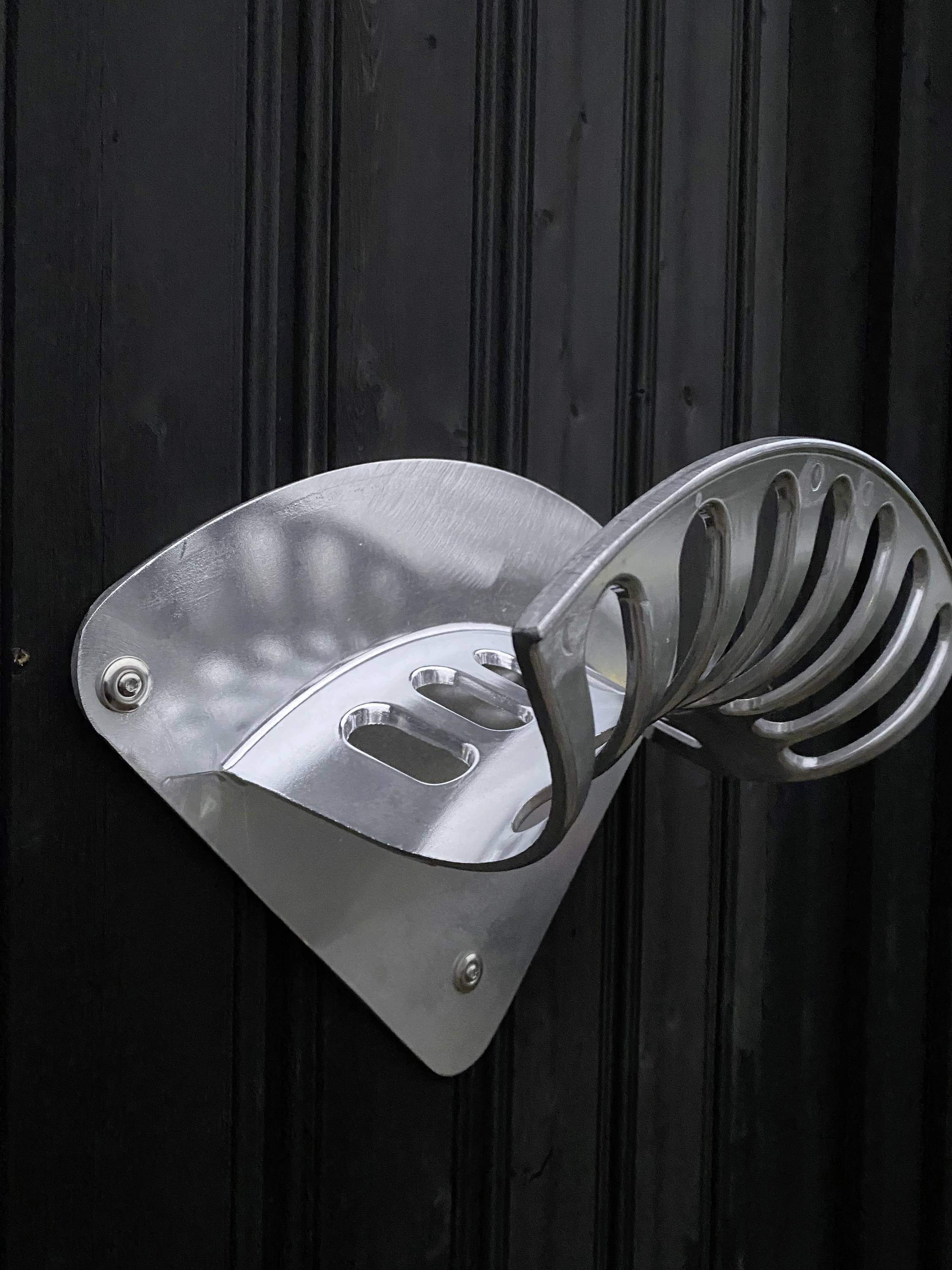 A silver, wall-mounted, metallic object with a curved design, likely a bicycle rack, against black wooden panelling.