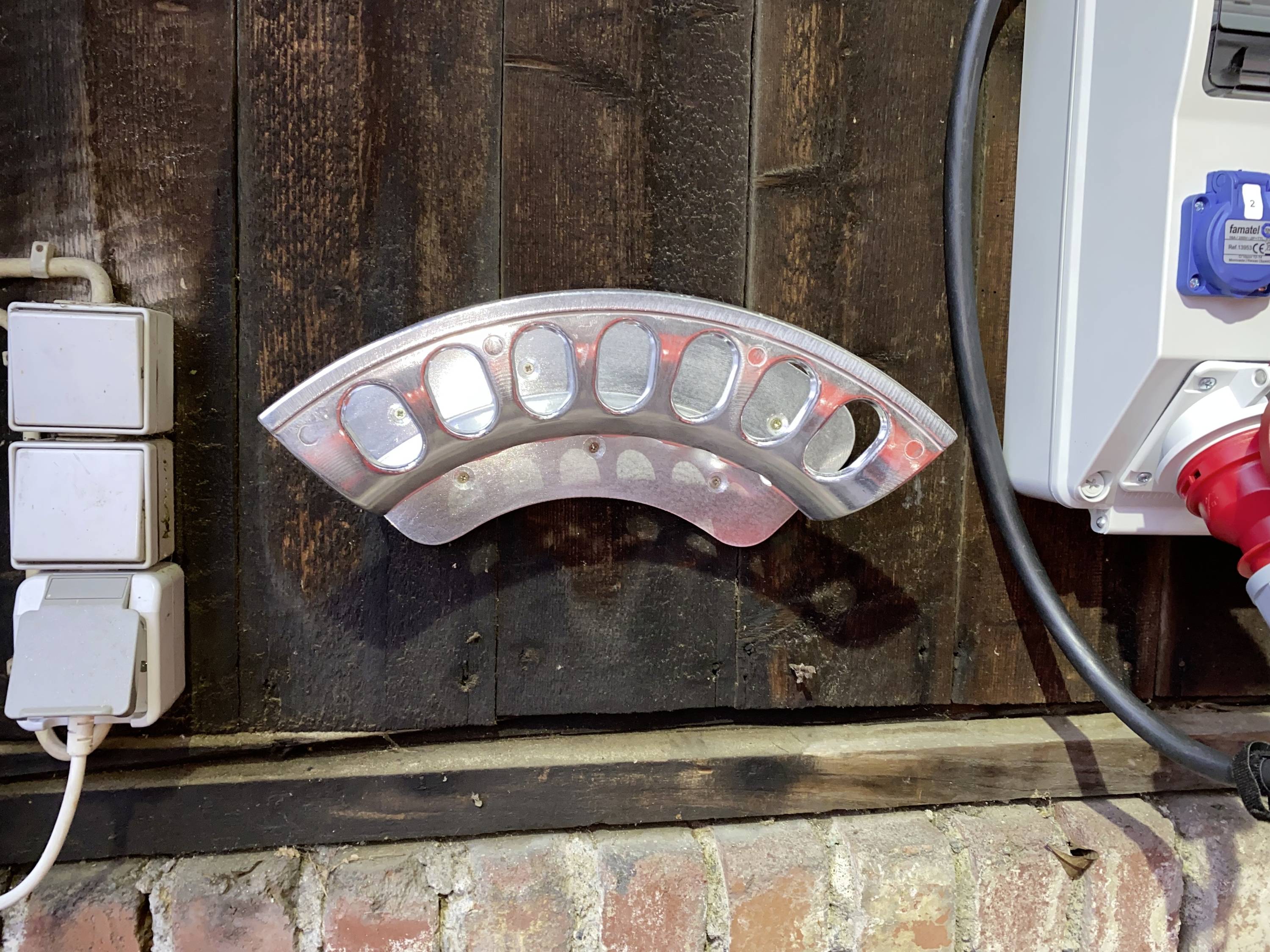 Metal bracket mounted on a wooden wall, surrounded by plug sockets and electrical installations, in front of a brick wall.