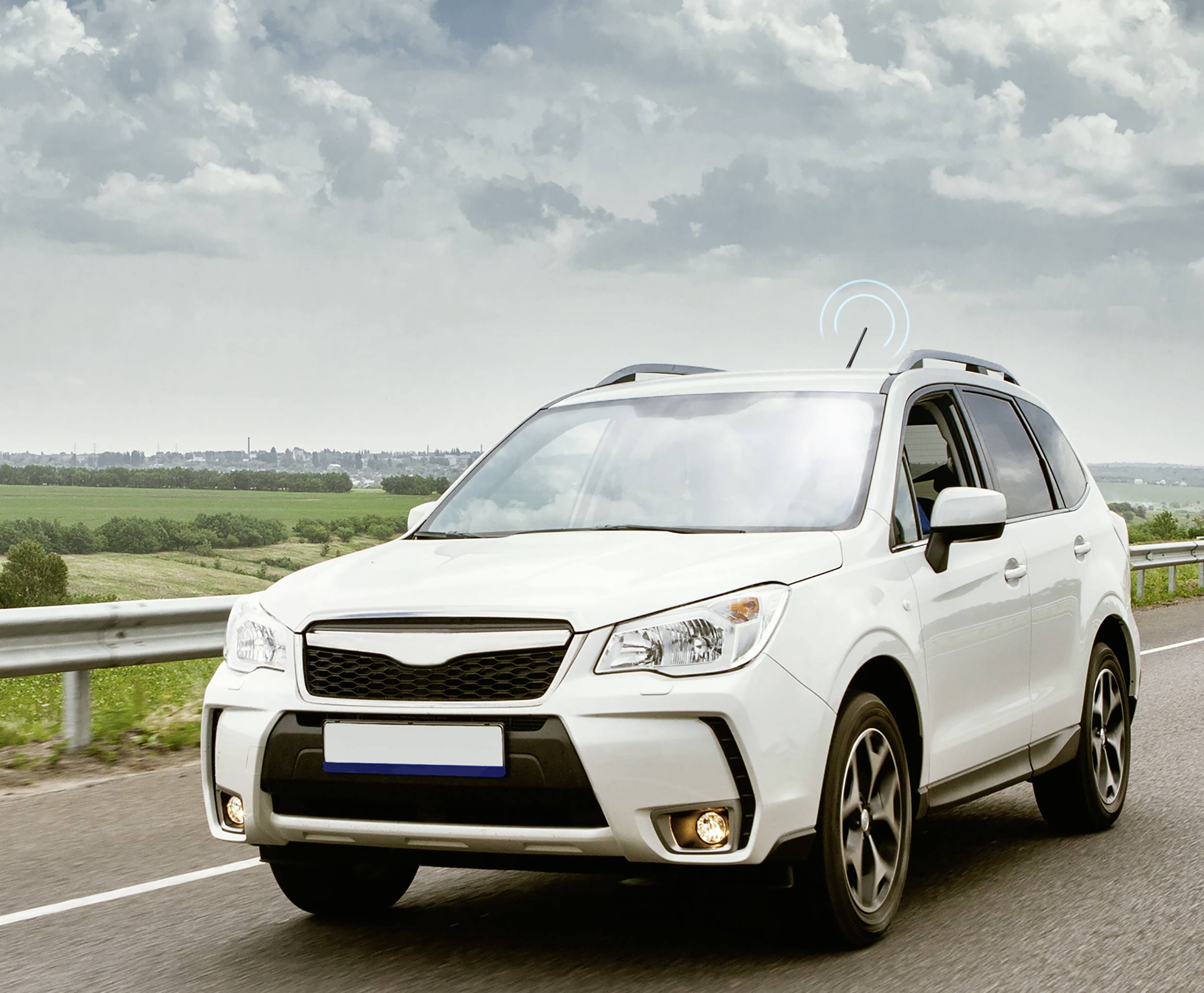 A white SUV is driving along a country road under a cloudy sky. Fields and a town can be seen in the background.