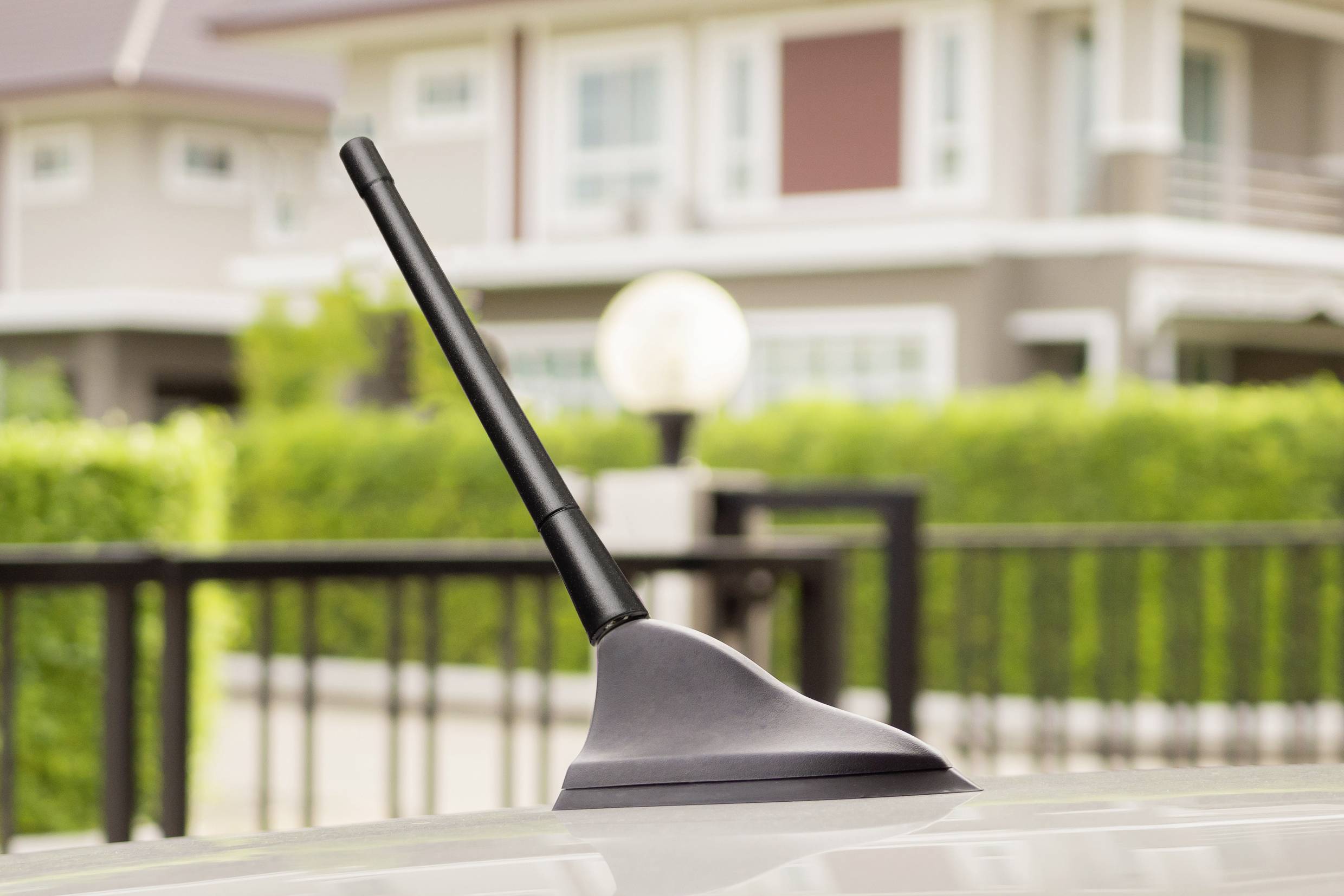 Car aerial mounted on a vehicle roof, with a blurred residential house and green garden in the background.