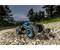 Remote-controlled car driving over a rocky path in rough terrain, with blue accents, surrounded by trees at dusk.