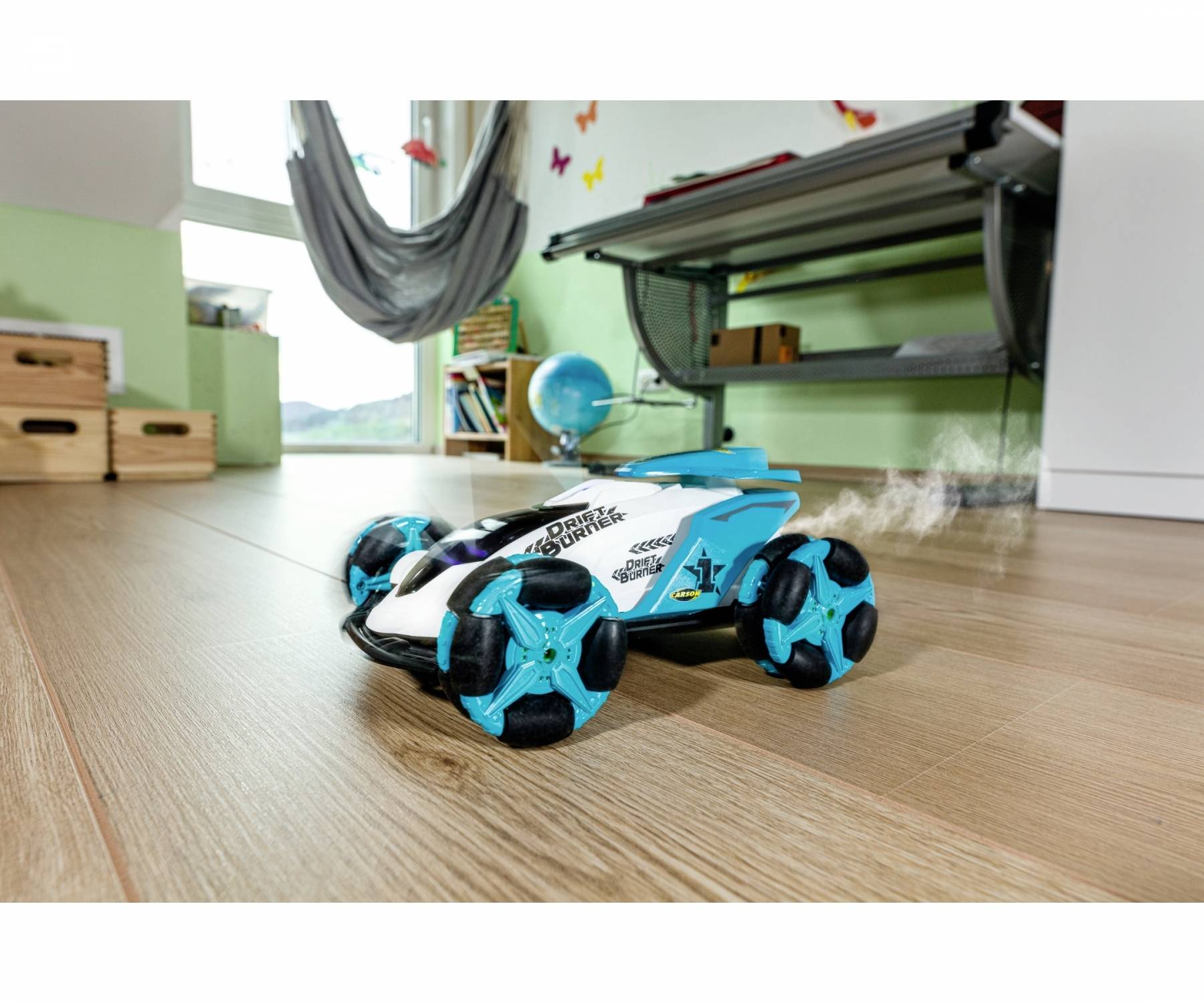 A blue and white remote-controlled car with large wheels drives across a wooden floor in a room. Toys and furniture are visible in the background.