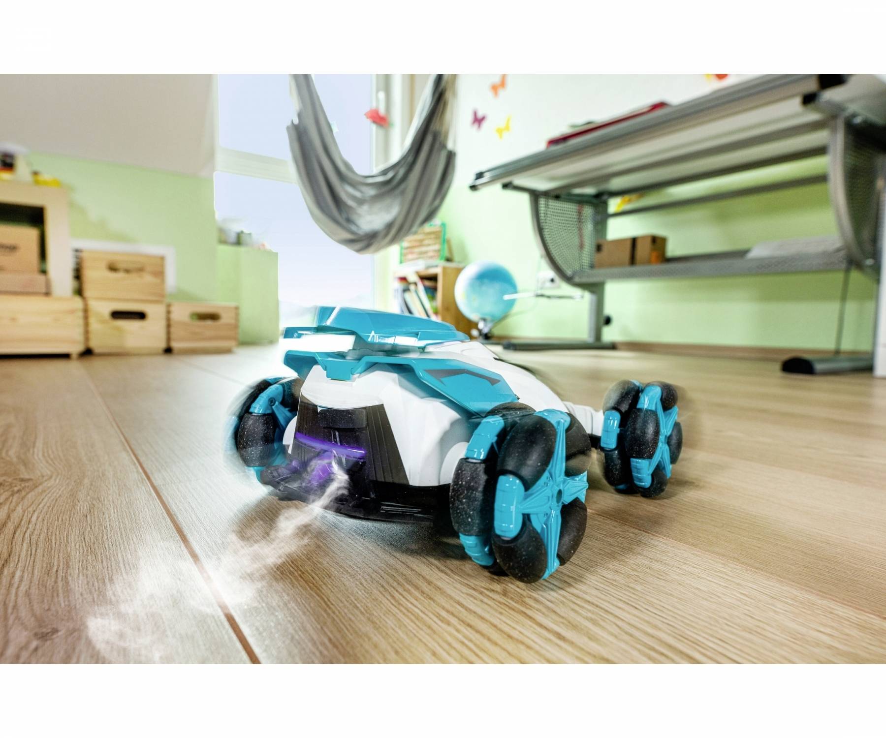 A toy car with blue wheels is driving on a wooden floor. In the background, a hanging chair, a table, and some boxes can be seen.