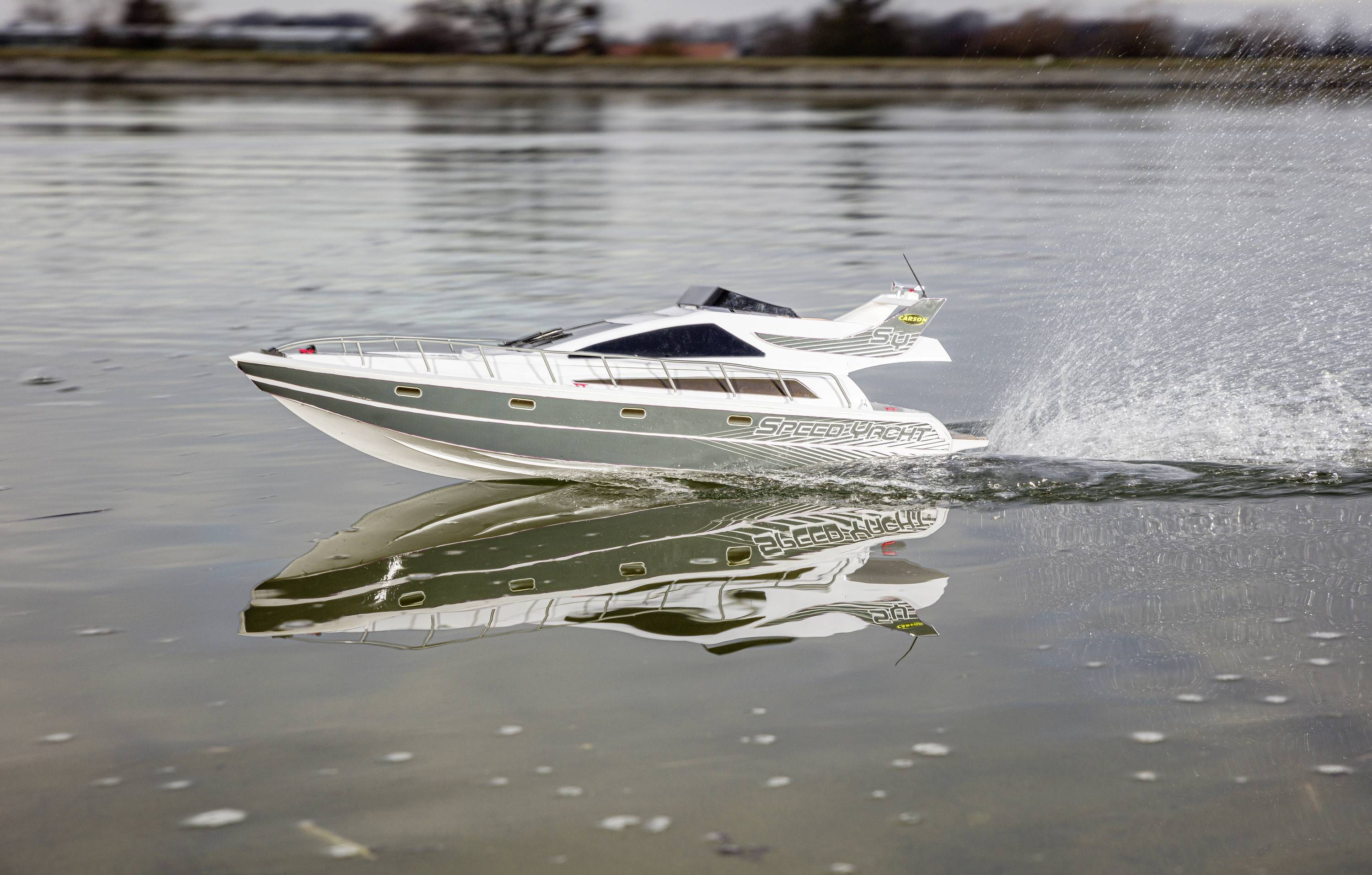 A remote-controlled model boat speeds across the water, spraying water and reflecting in the water's surface.
