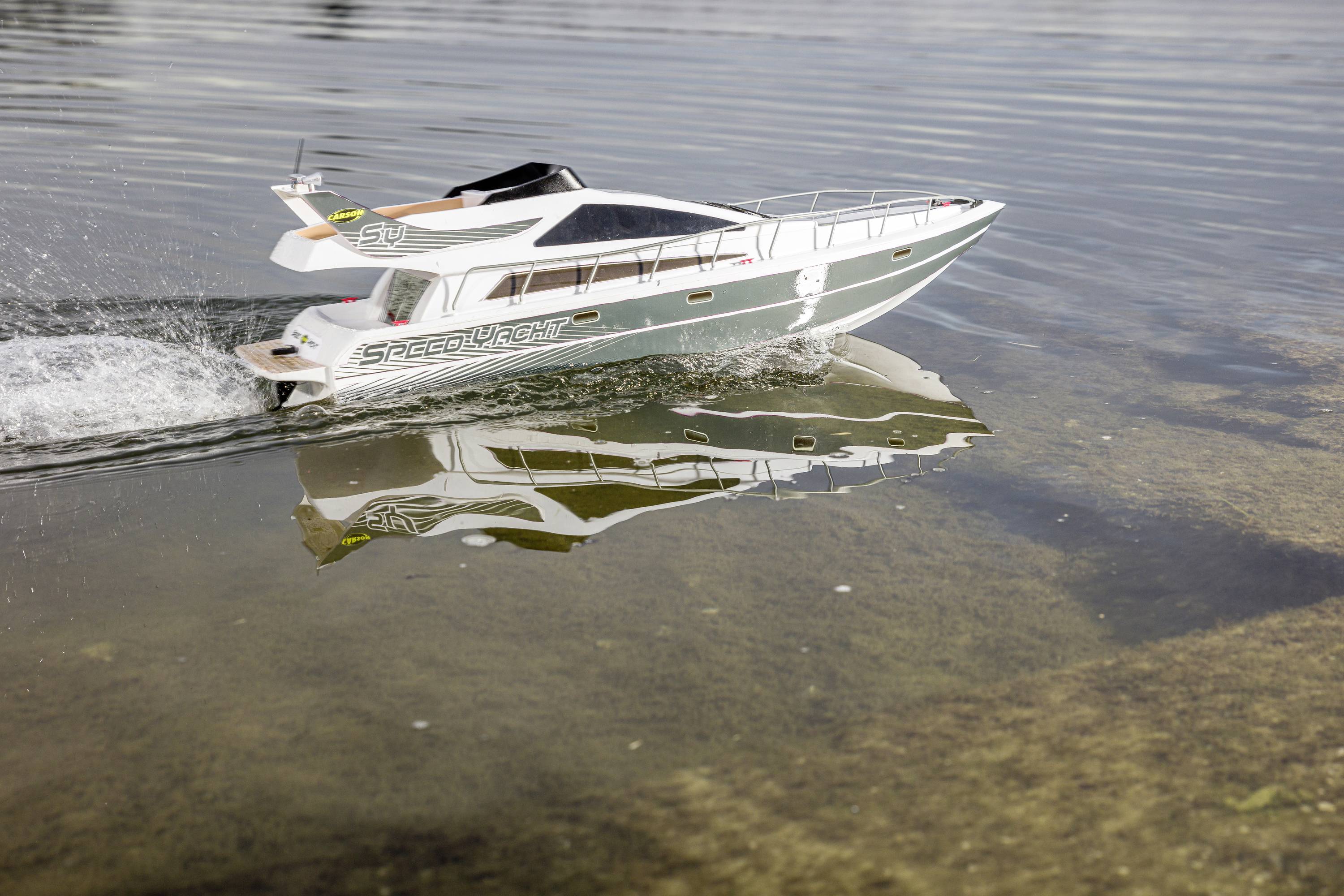 A remote-controlled model boat sails on a calm, flat body of water. The boat splashes water and is reflected in the water.