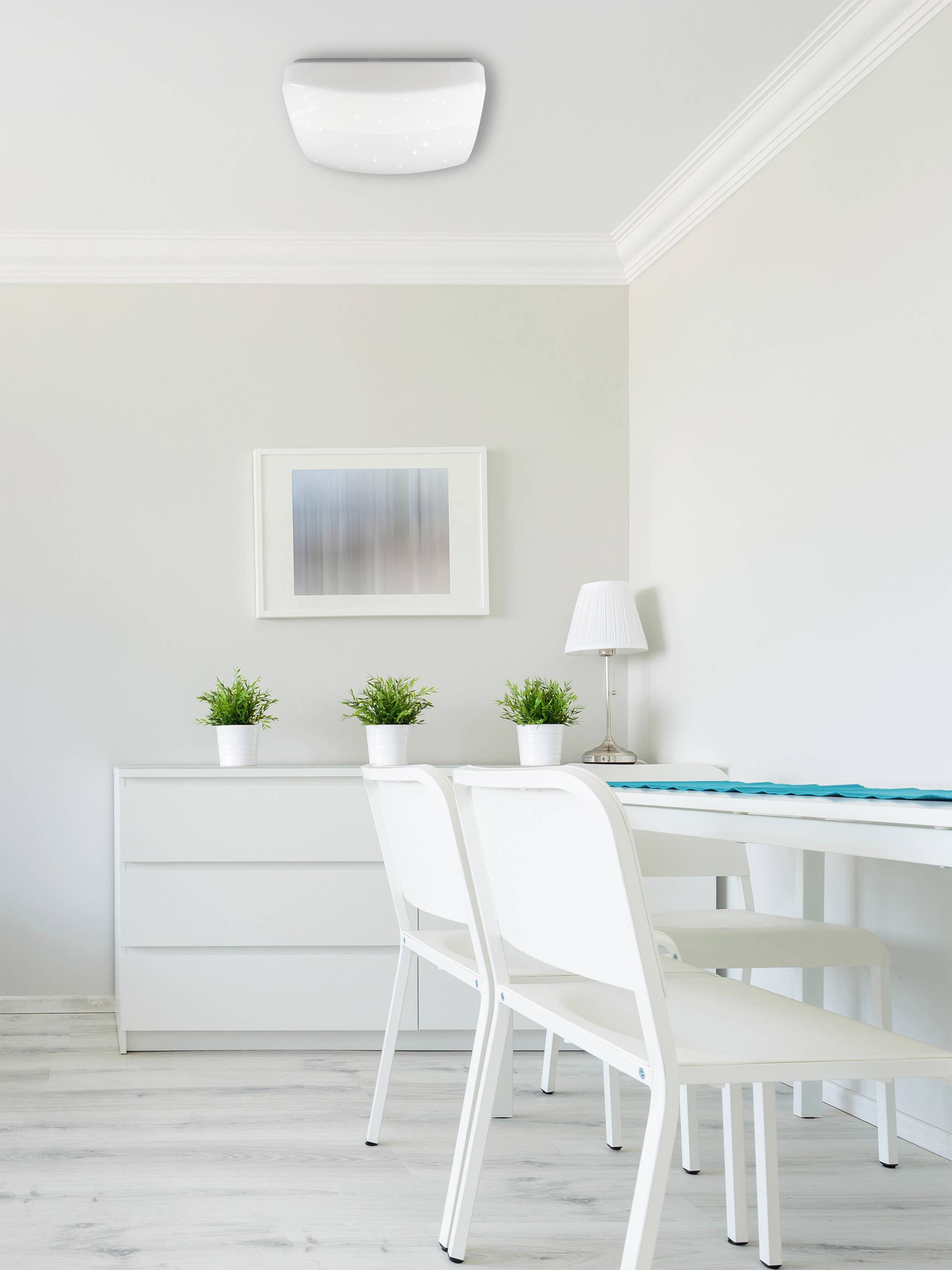 A bright, modern room with a white table, two chairs, and green plants on a sideboard. A framed picture hangs on the wall.
