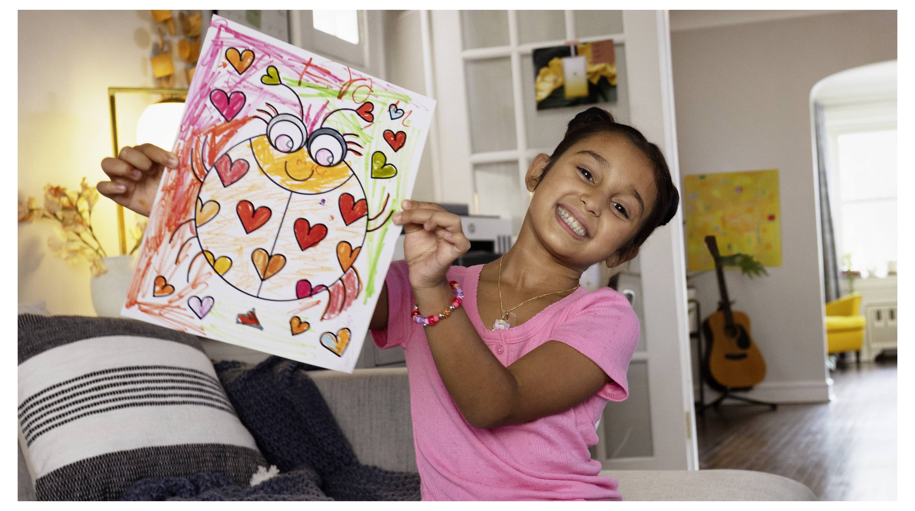 Smiling girl in a pink shirt shows a colorful heart-covered drawing to the camera in a home living room.