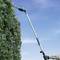 A person is trimming a hedge with long hedge shears outdoors. The sky is clear, and it is daytime.