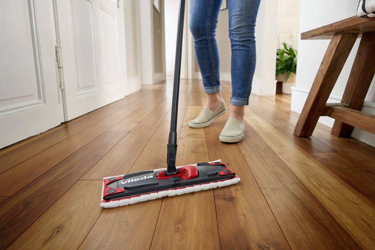 A person is mopping the wooden floor with a rectangular mop. A light-coloured hallway and doors are visible in the background.