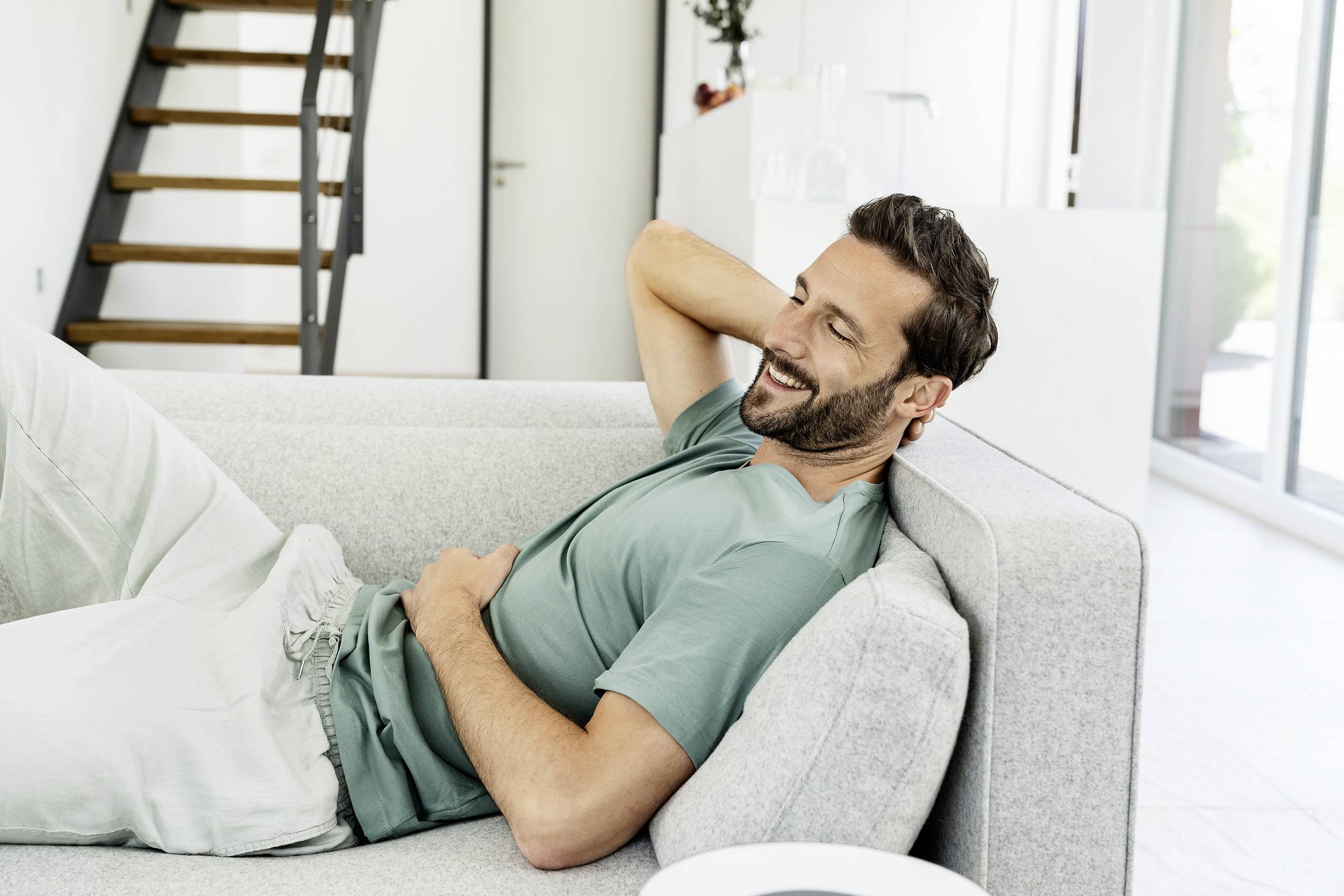 A man is lying relaxed on a grey sofa, smiling with his hands clasped behind his head. A staircase can be seen in the background.