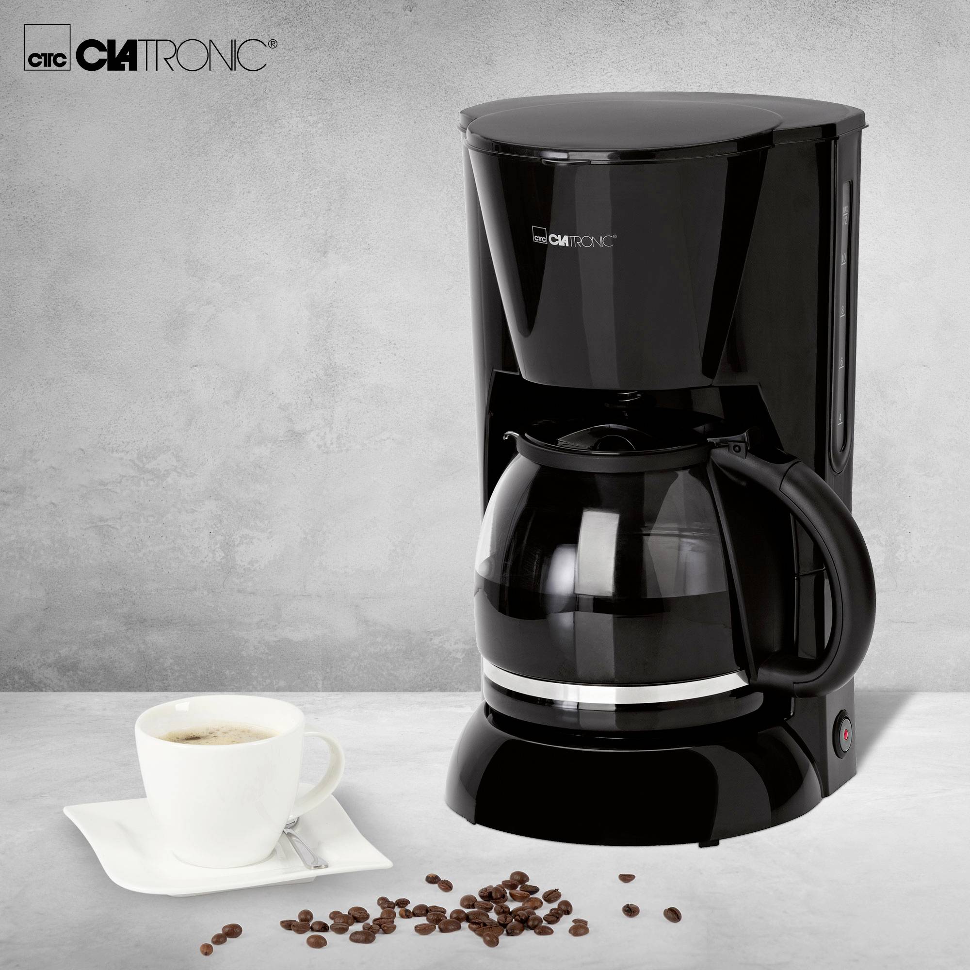 Coffee machine with glass carafe on grey background. Beside it stands a cup of coffee. Coffee beans are scattered.
