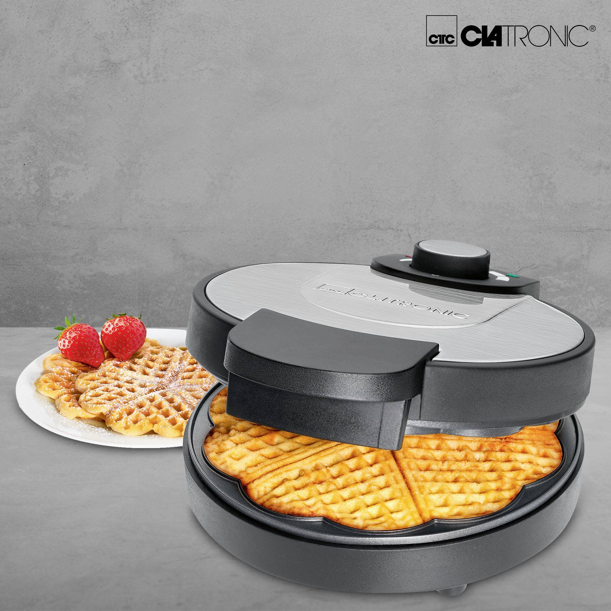 Waffle iron with baked waffles, alongside a plate of finished waffles and strawberries.
