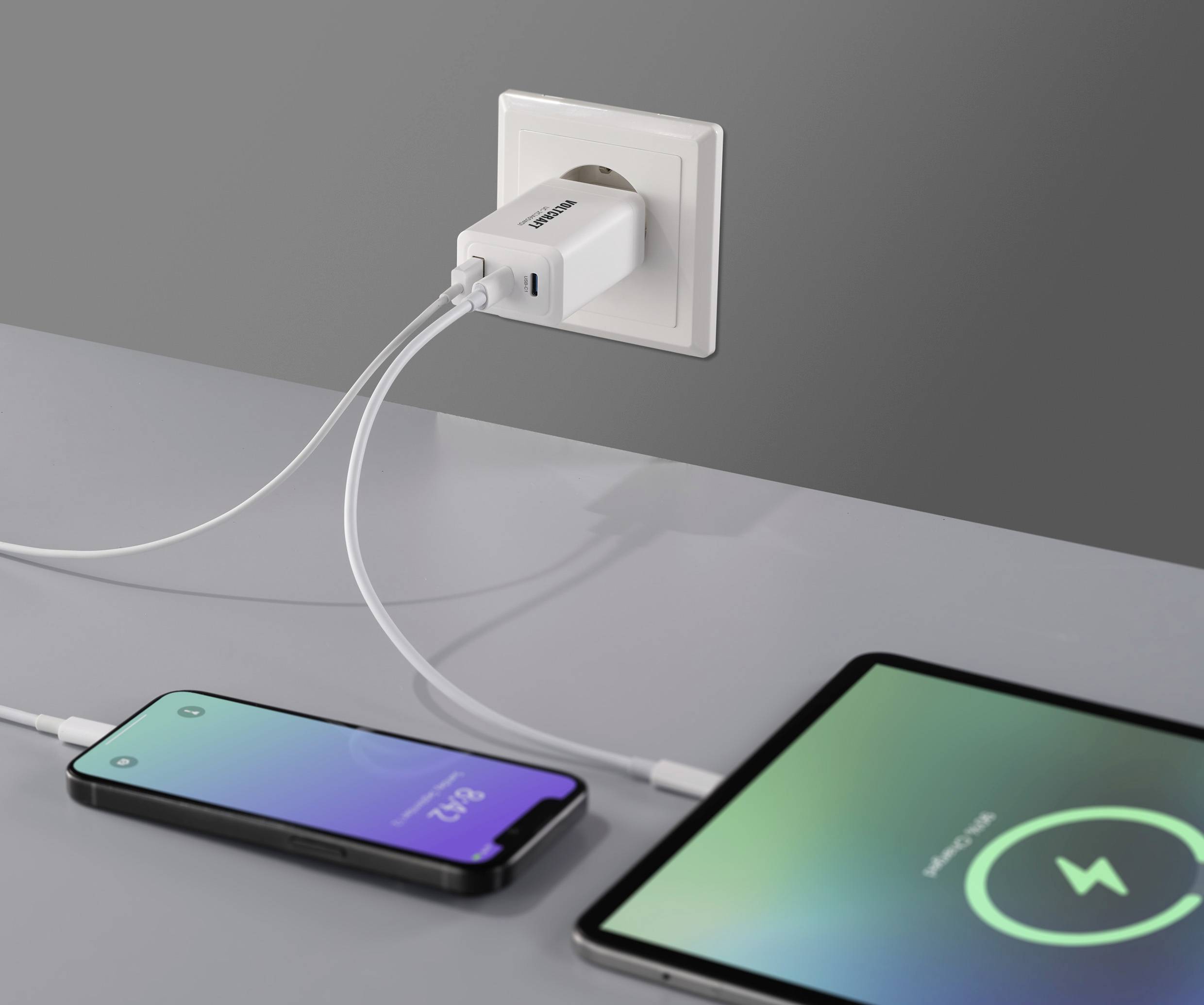 A charging adapter is plugged into a power socket and is simultaneously charging a smartphone and a tablet. Both devices are displaying charging screens.