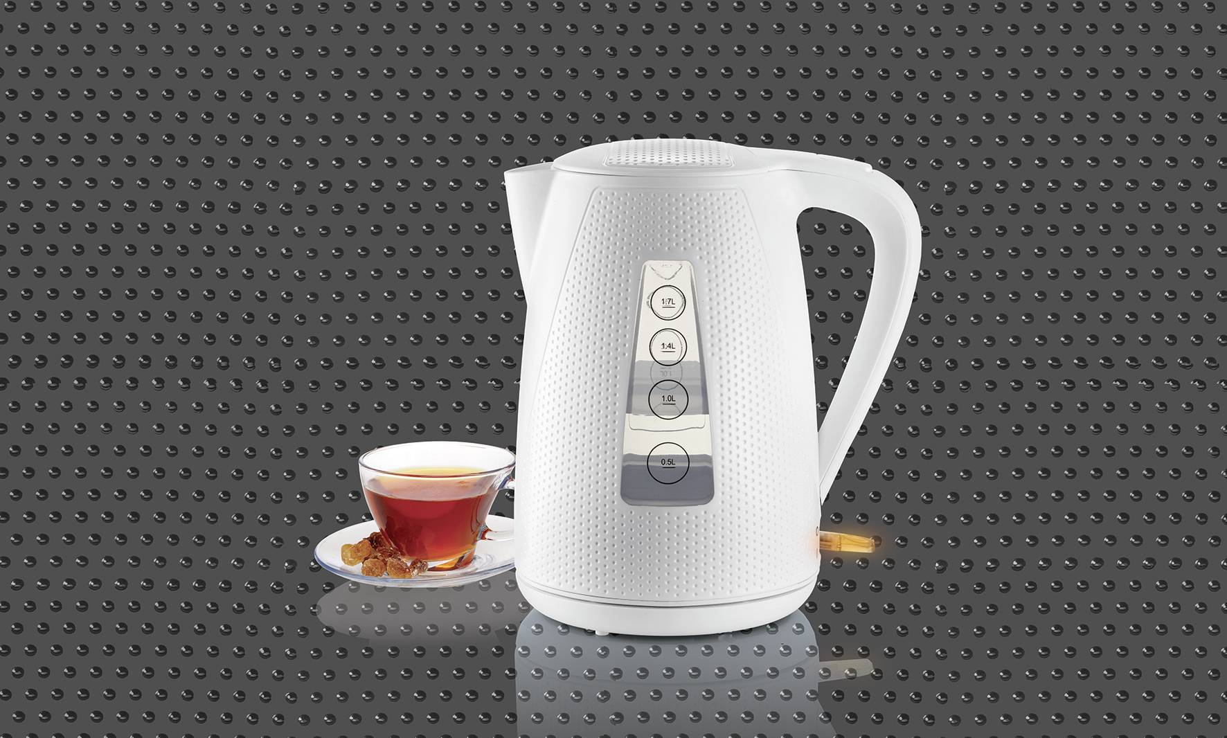 A white kettle sits against a dark background with a glass of tea on a saucer beside it.