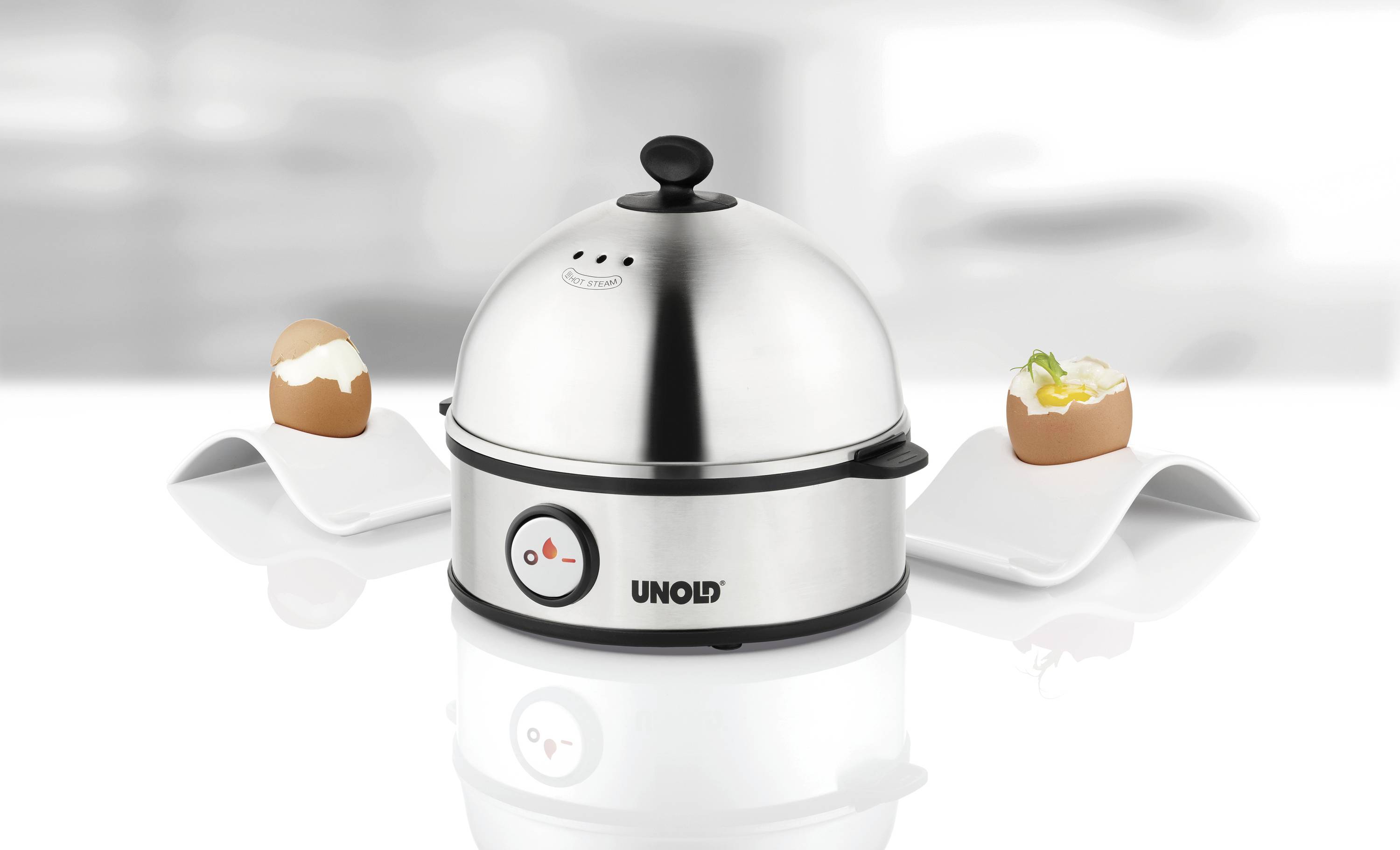 A silver egg cooker with a black handle from the UNOLD brand, surrounded by two halved, prepared eggs on white plates.