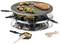 A raclette grill with a hot stone, on which meat, vegetables, and mushrooms are sizzling. Underneath, small pans with cheese and wooden spatulas.