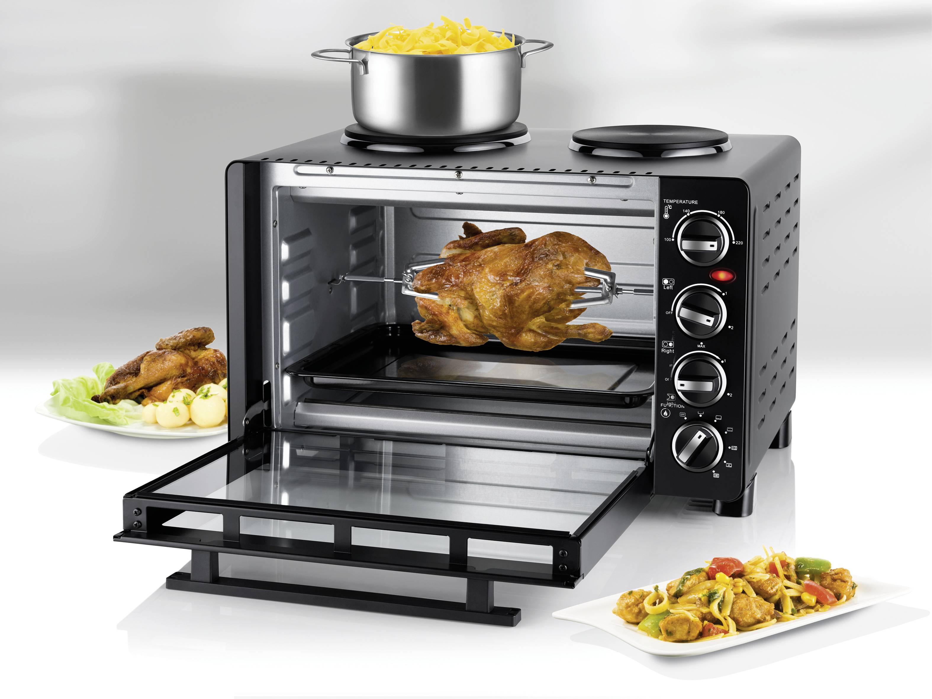 An electric oven with an open door shows a roast on a spit. A pot of pasta sits on the oven. Plates with food are placed to the left and right.