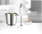 An immersion blender next to a pot of soup and a bowl on a kitchen worktop, with a blurry kitchen in the background.