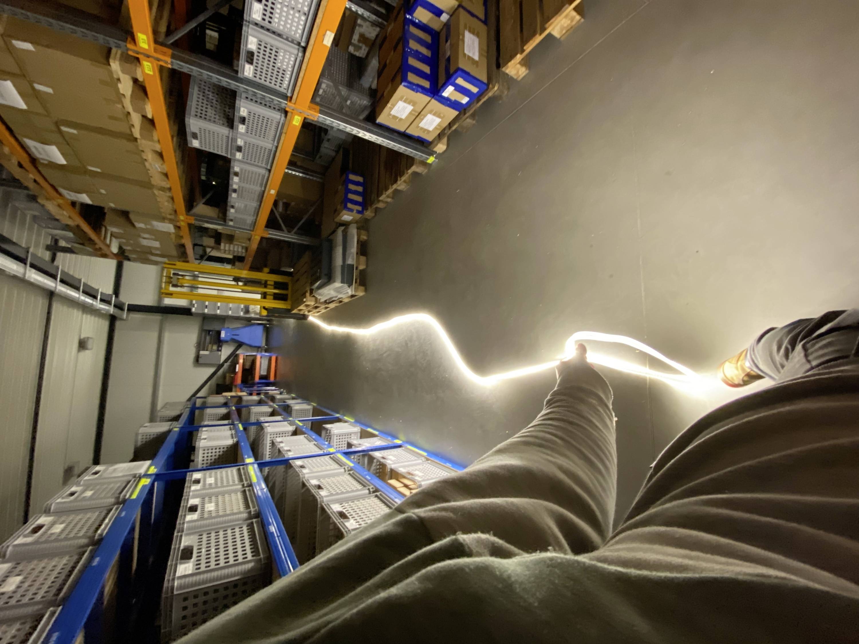 A person in a warehouse pulls a glowing cable along a corridor lined with shelves stacked with boxes. A bright, undulating path of light is created.