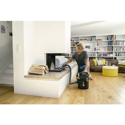 A person is using an ash vacuum to clean a fireplace in a modern living room with a wooden floor and bookshelf.