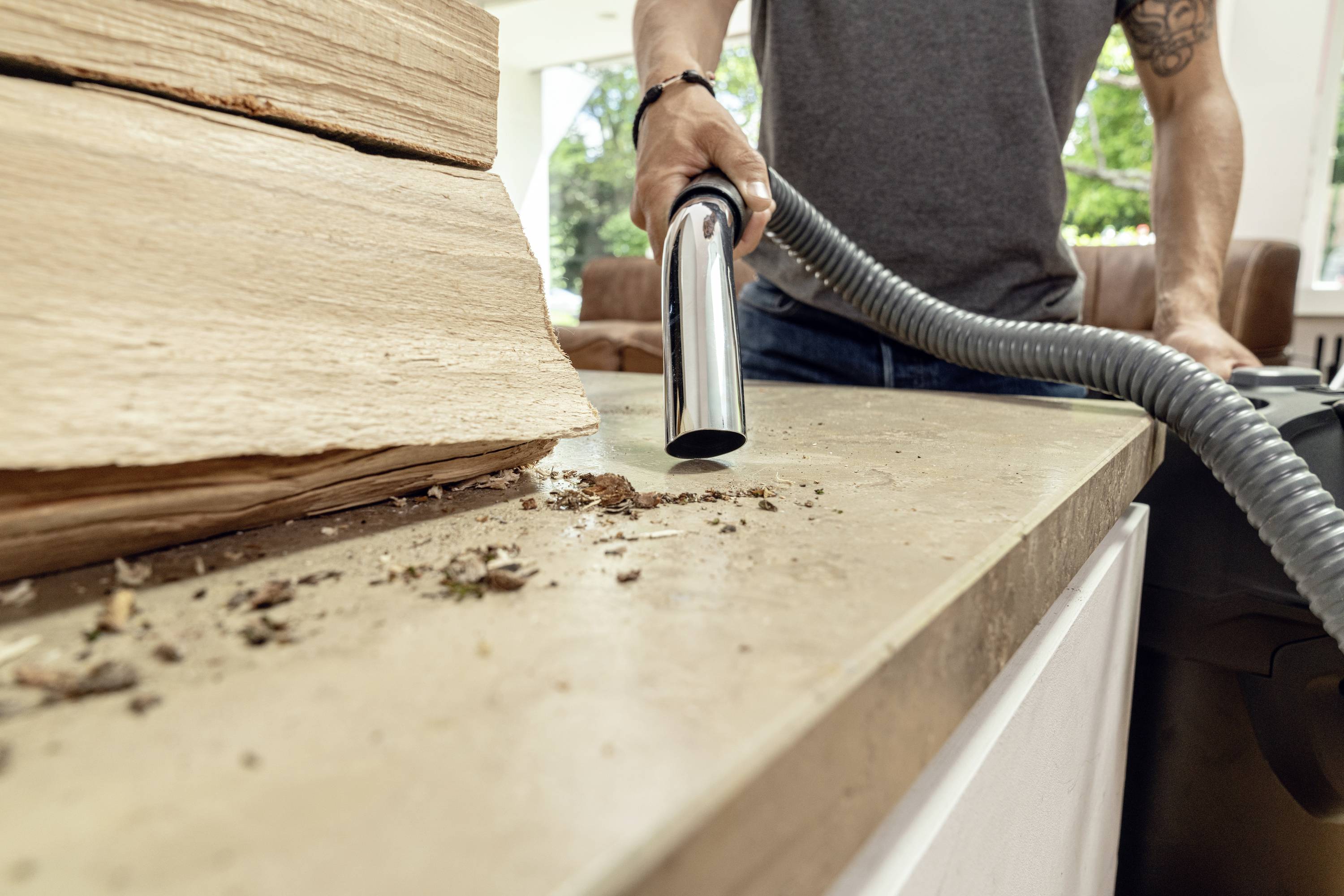 A person is vacuuming sawdust and dirt from a workbench. Trees can be seen in the background.