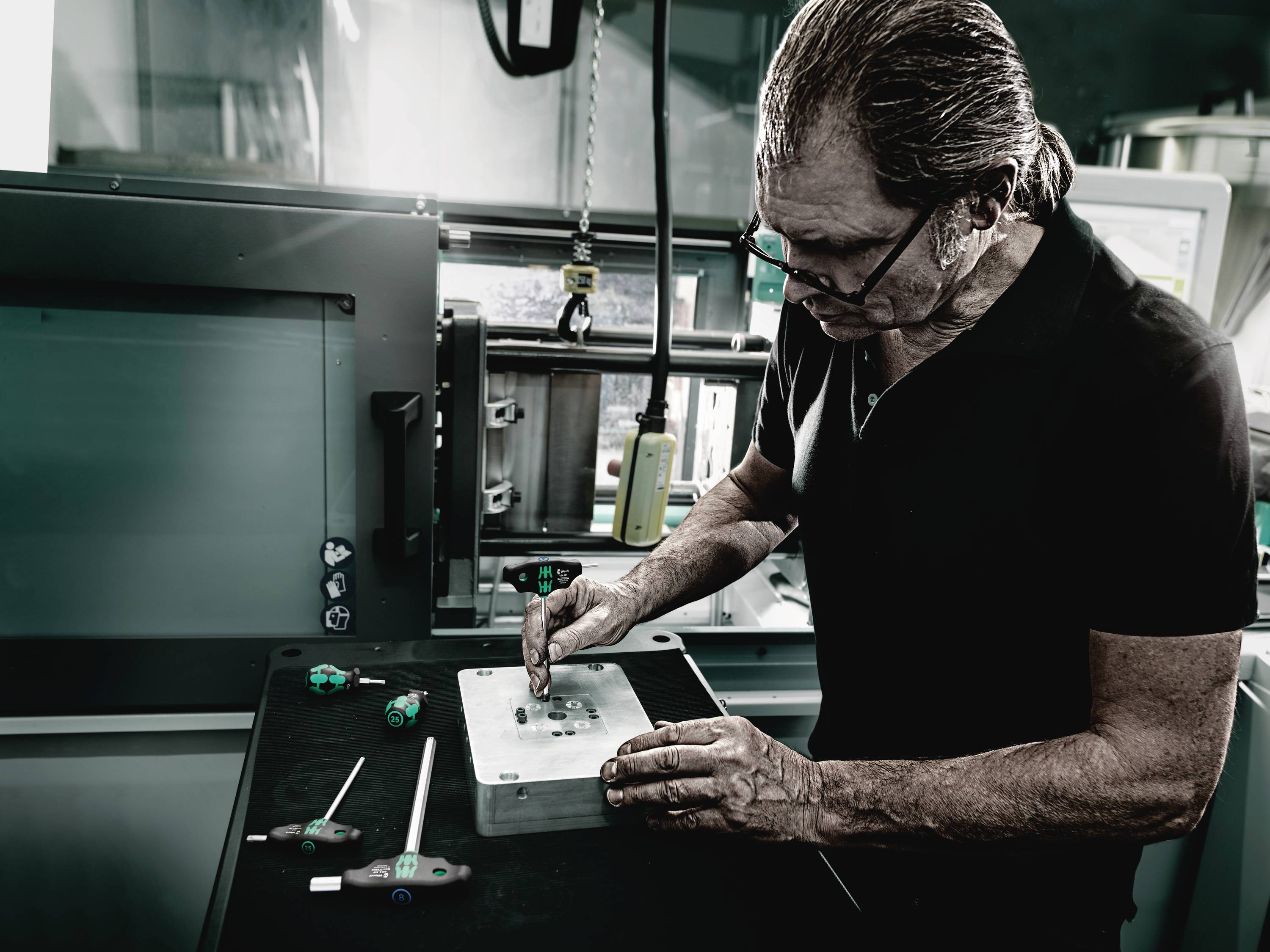 An older man is working intently on a metal block in a workshop. Tools are scattered around him.