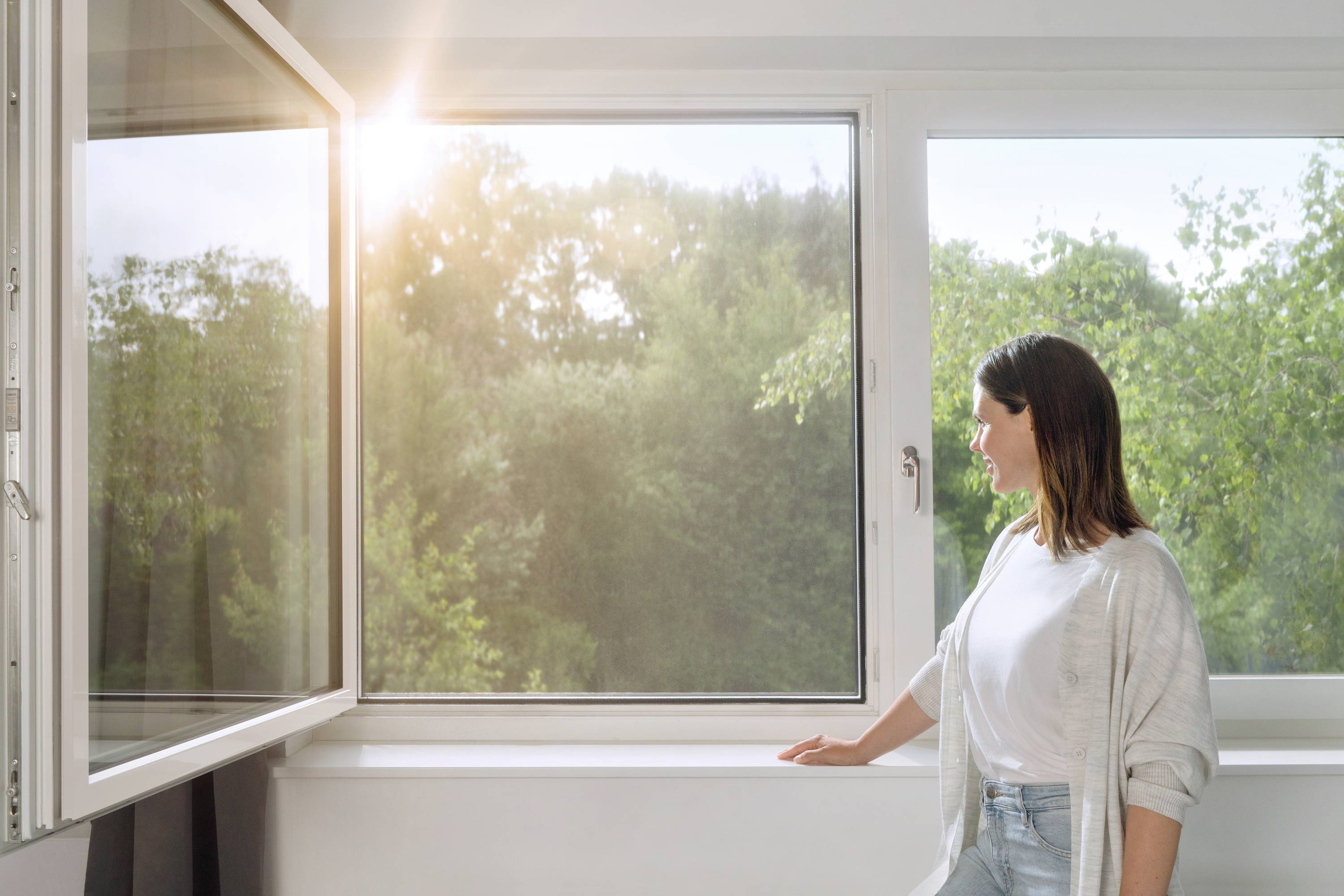 A woman stands by an open window, gazing outside, while sunlight illuminates the room. Green trees are visible outside.