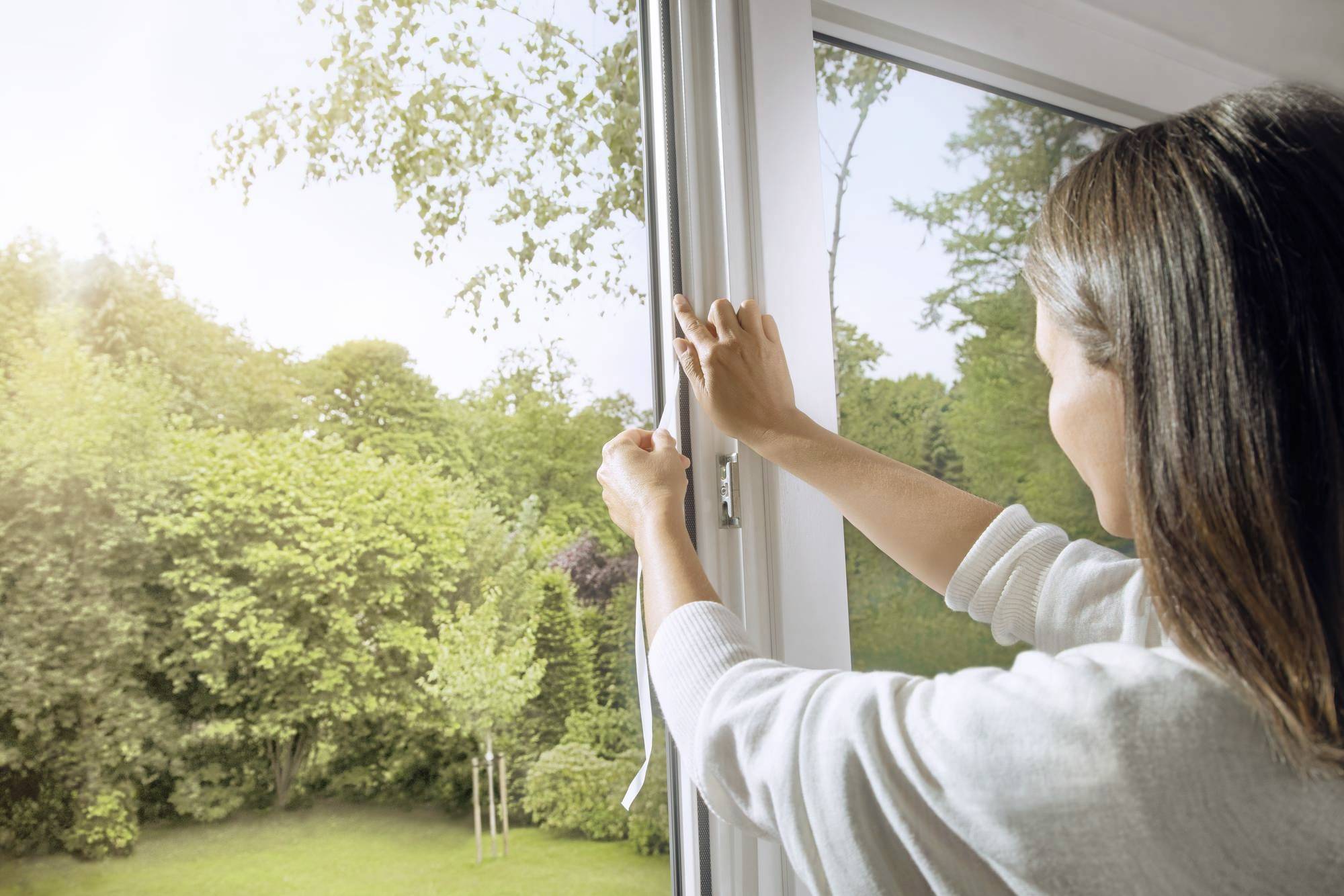 A woman opens a window, through which a sunny garden with trees is visible.