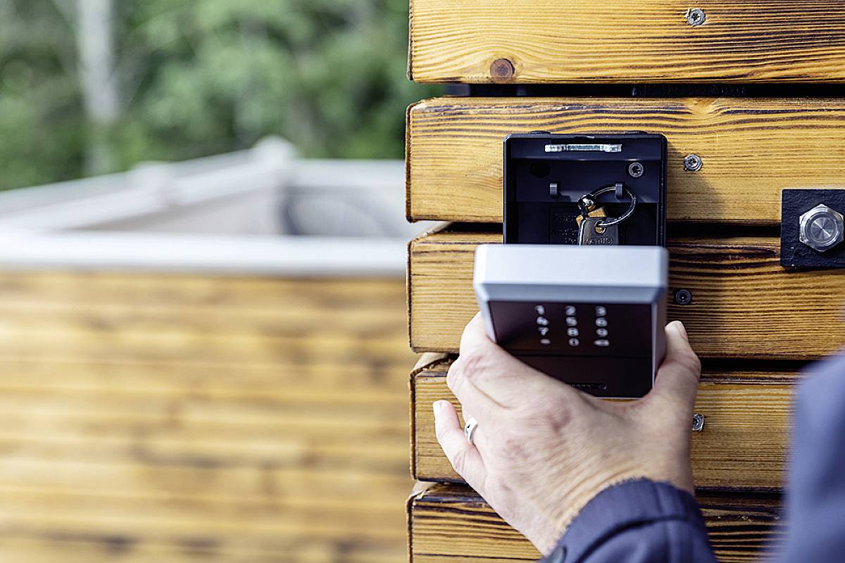 A person is holding a numerical lock device mounted on a wooden wall. A blurry, grass-covered area is visible in the background.