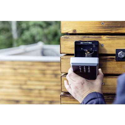 A person is holding a numerical lock device mounted on a wooden wall. A blurry, grass-covered area is visible in the background.