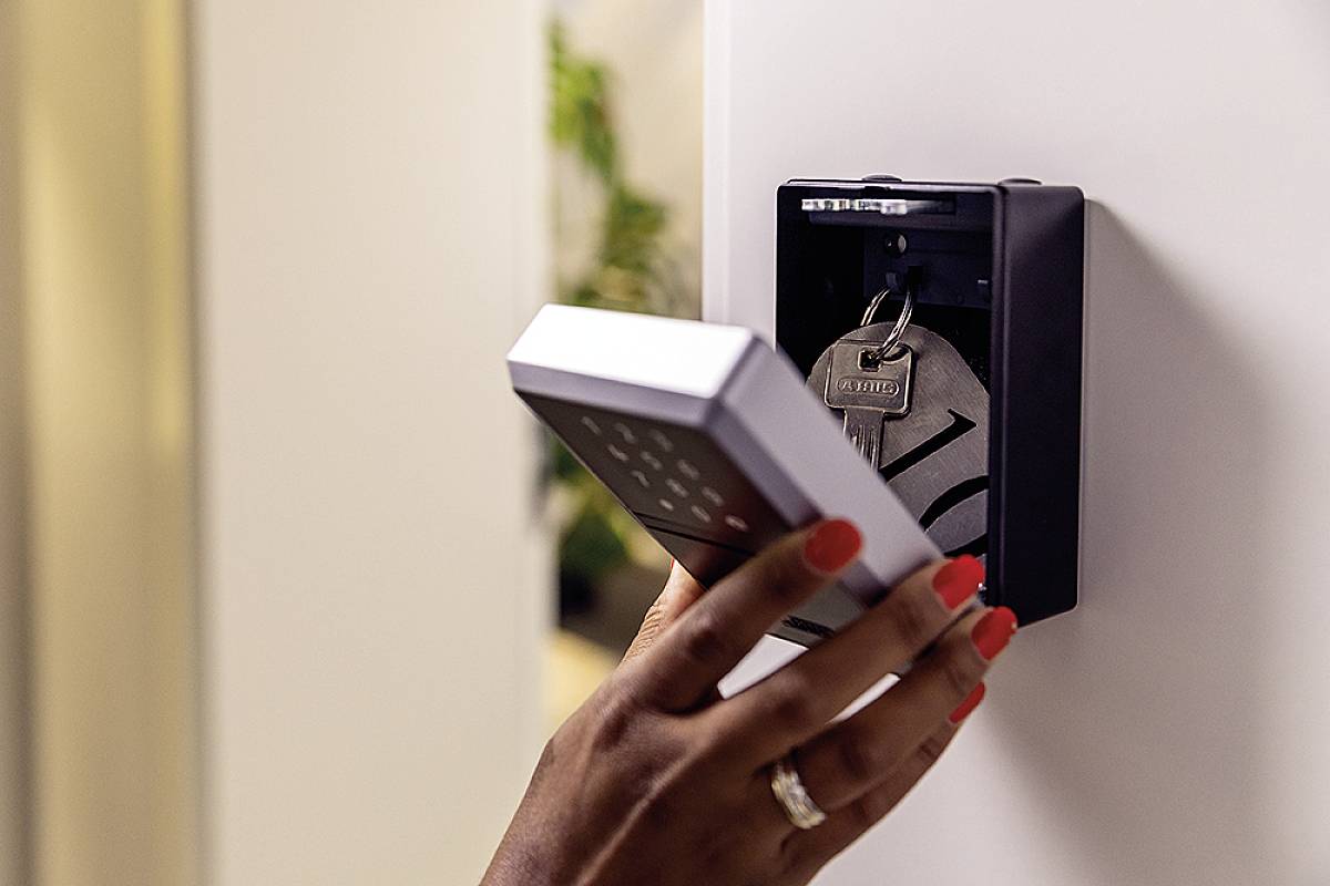 A hand opens an electronic door lock with a number keypad, revealing a key safe containing a bunch of keys.