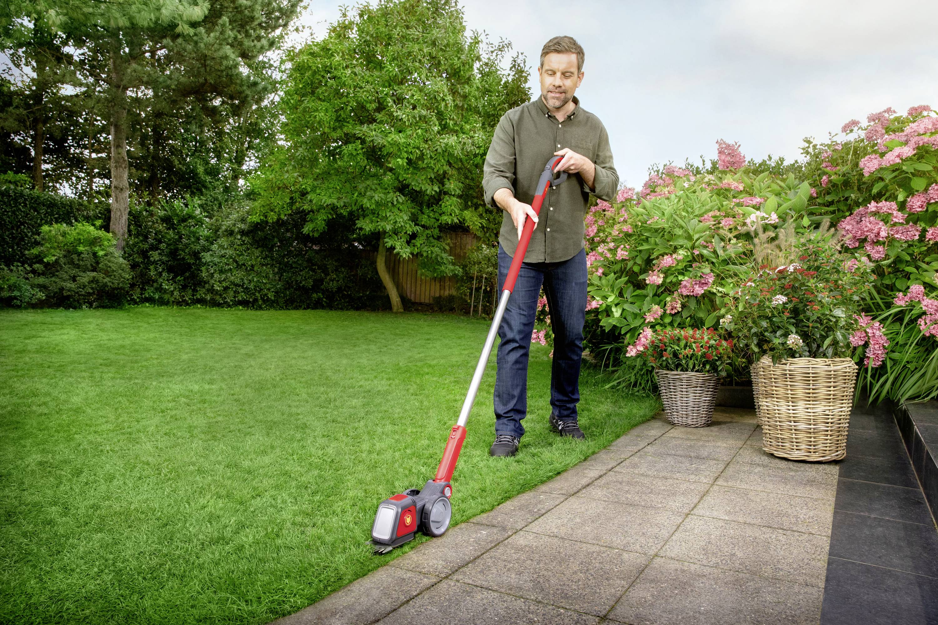 A man is using a lawn trimmer to edge a lawn next to flowering plants in baskets. Garden in the background.