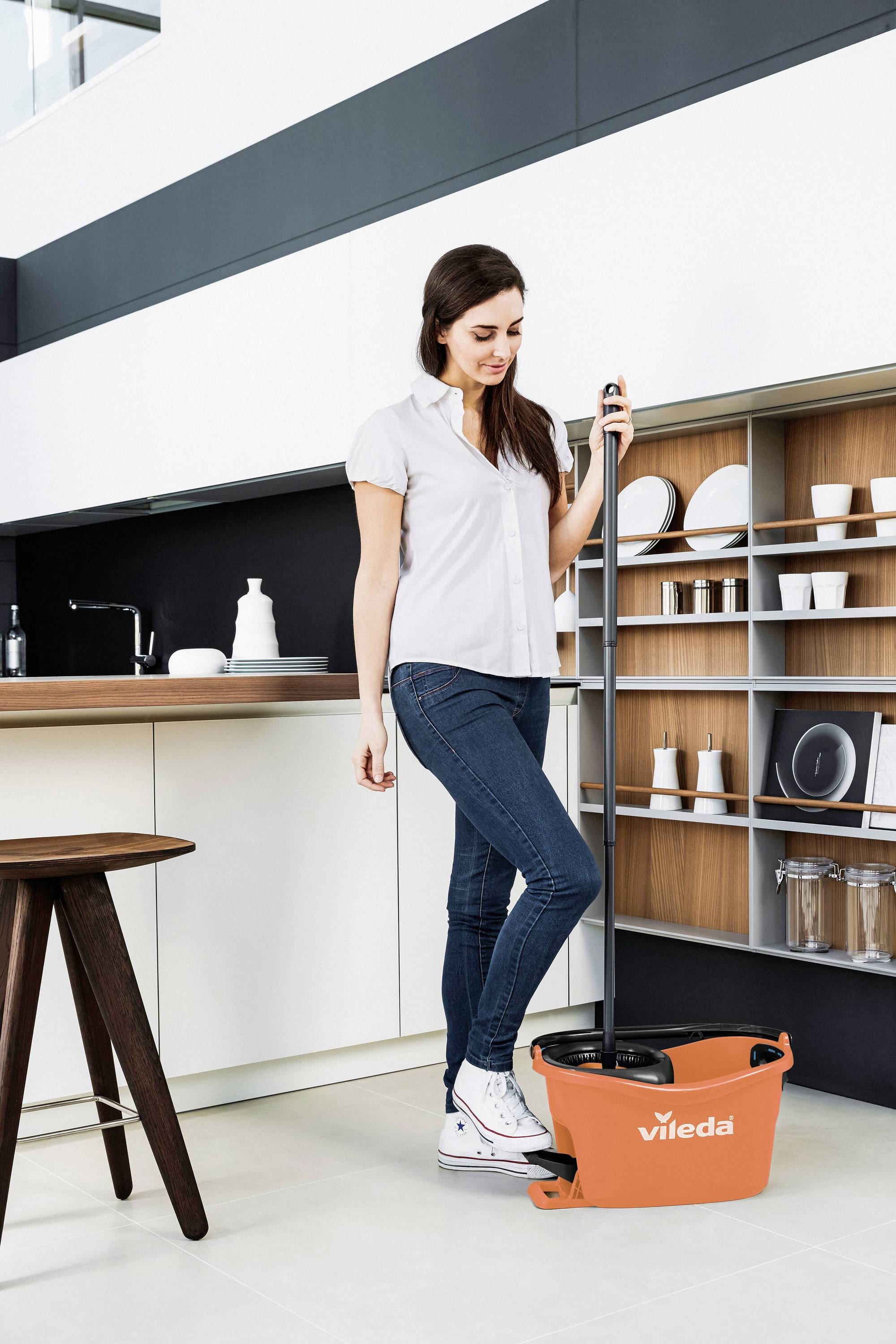 A woman in a kitchen is using a mop with an orange Vileda bucket. In the background, there is a shelf with crockery and a kitchen unit.