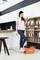 A woman in a kitchen is using a mop with an orange Vileda bucket. In the background, there is a shelf with crockery and a kitchen unit.