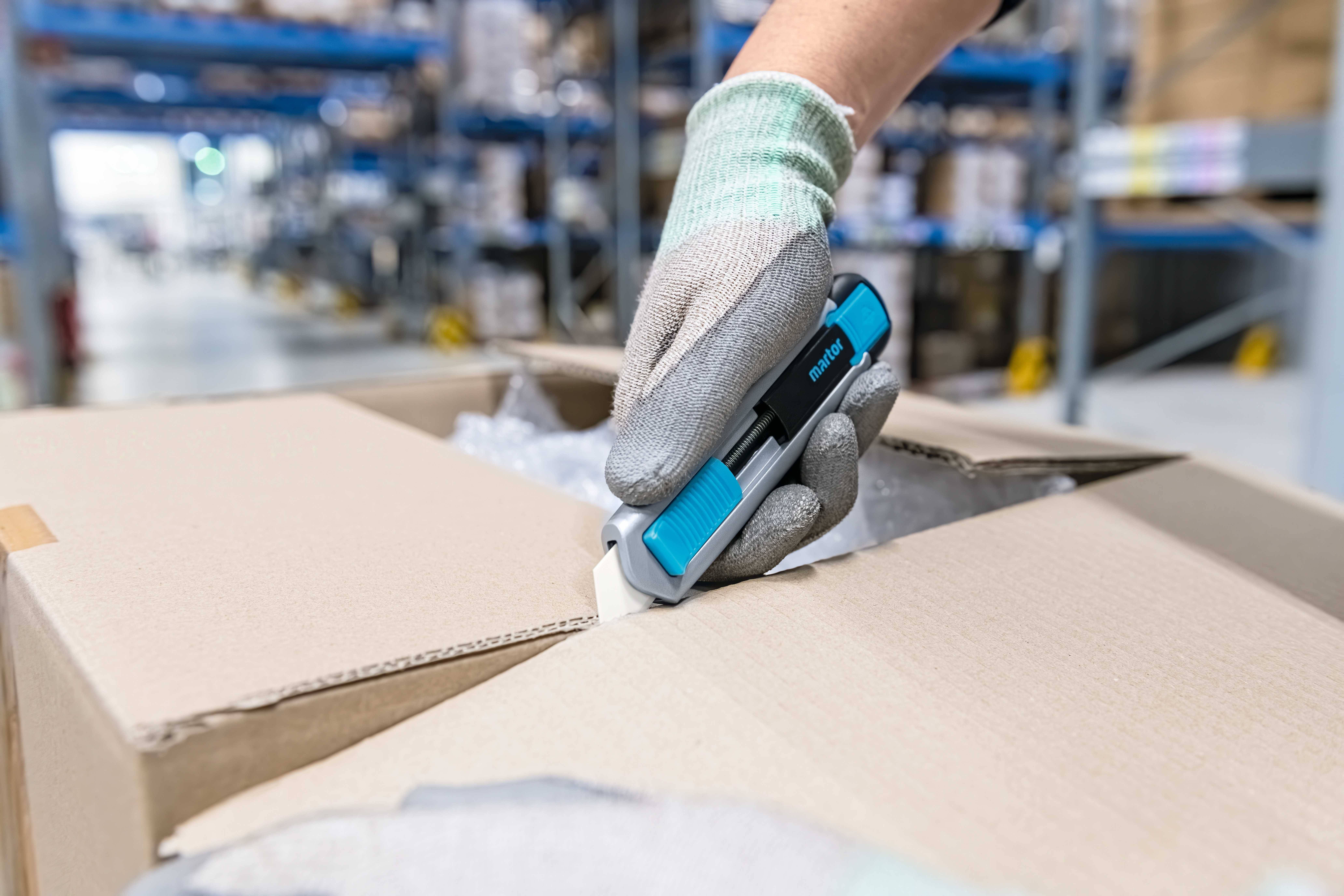 A person opens a cardboard box with a blue safety knife in a warehouse. Shelves of goods can be seen in the background.