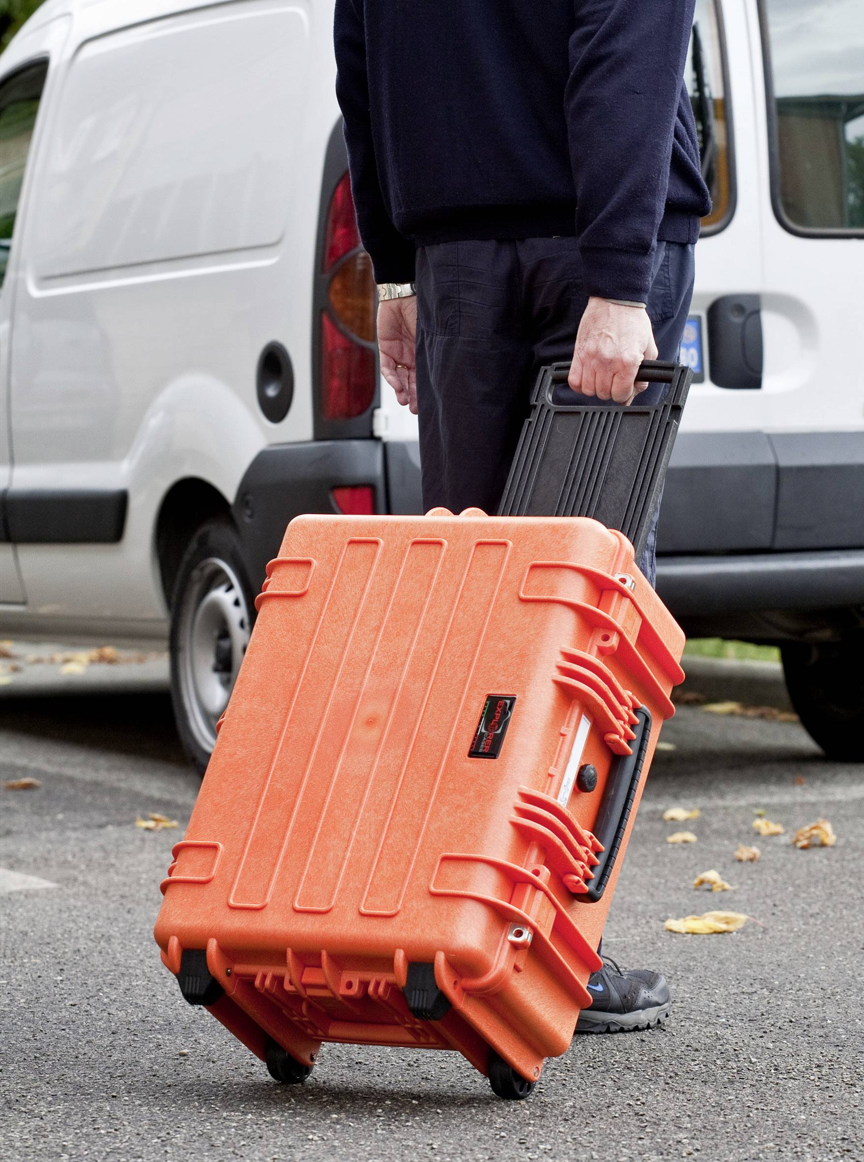 A person is pulling a large, orange suitcase with wheels across a car park next to a white vehicle.
