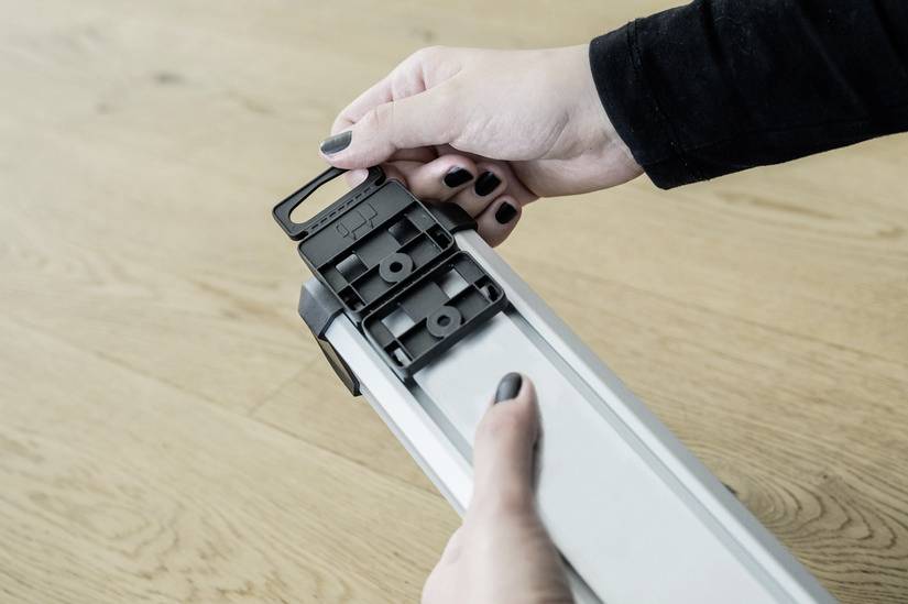 A person is holding a rectangular, silver box with a black fastener on a wooden floor and is opening it.