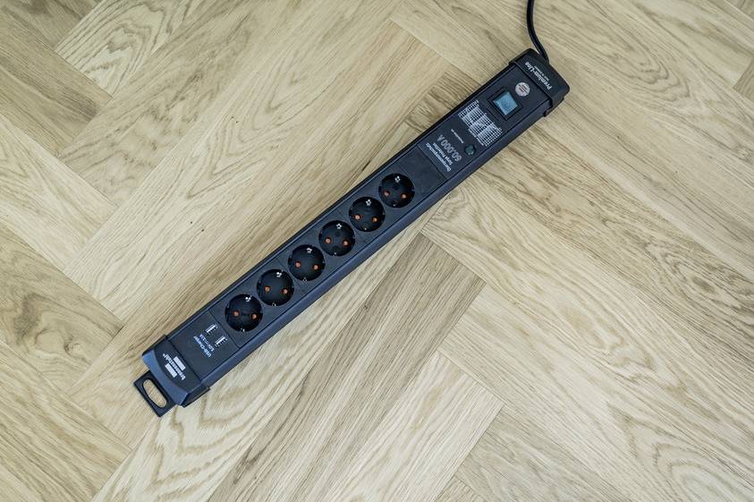A black multi-socket extension with five plug sockets and a switch is lying on a light wooden floor.