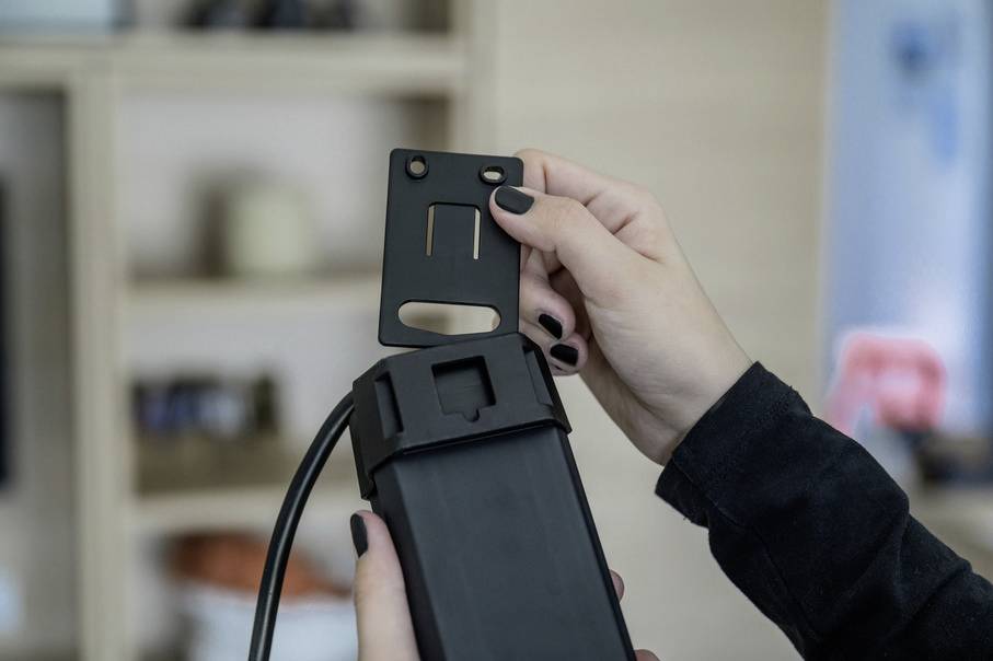 A person is holding a black electronic device in their hand and attaching a cover to it. A shelf is visible in the background.