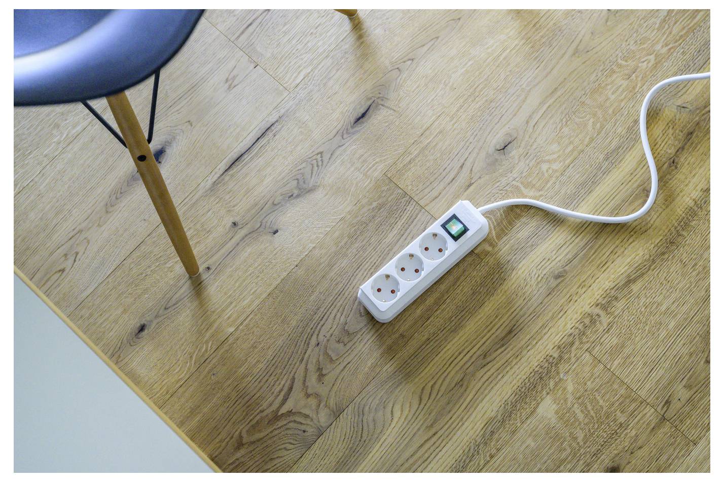 A white power strip with three outlets and a green switch lies on a wooden floor beside a chair leg, with its cord trailing off frame.