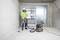 A construction worker in protective clothing is cleaning an empty room with an industrial vacuum cleaner. Large windows are in the background.