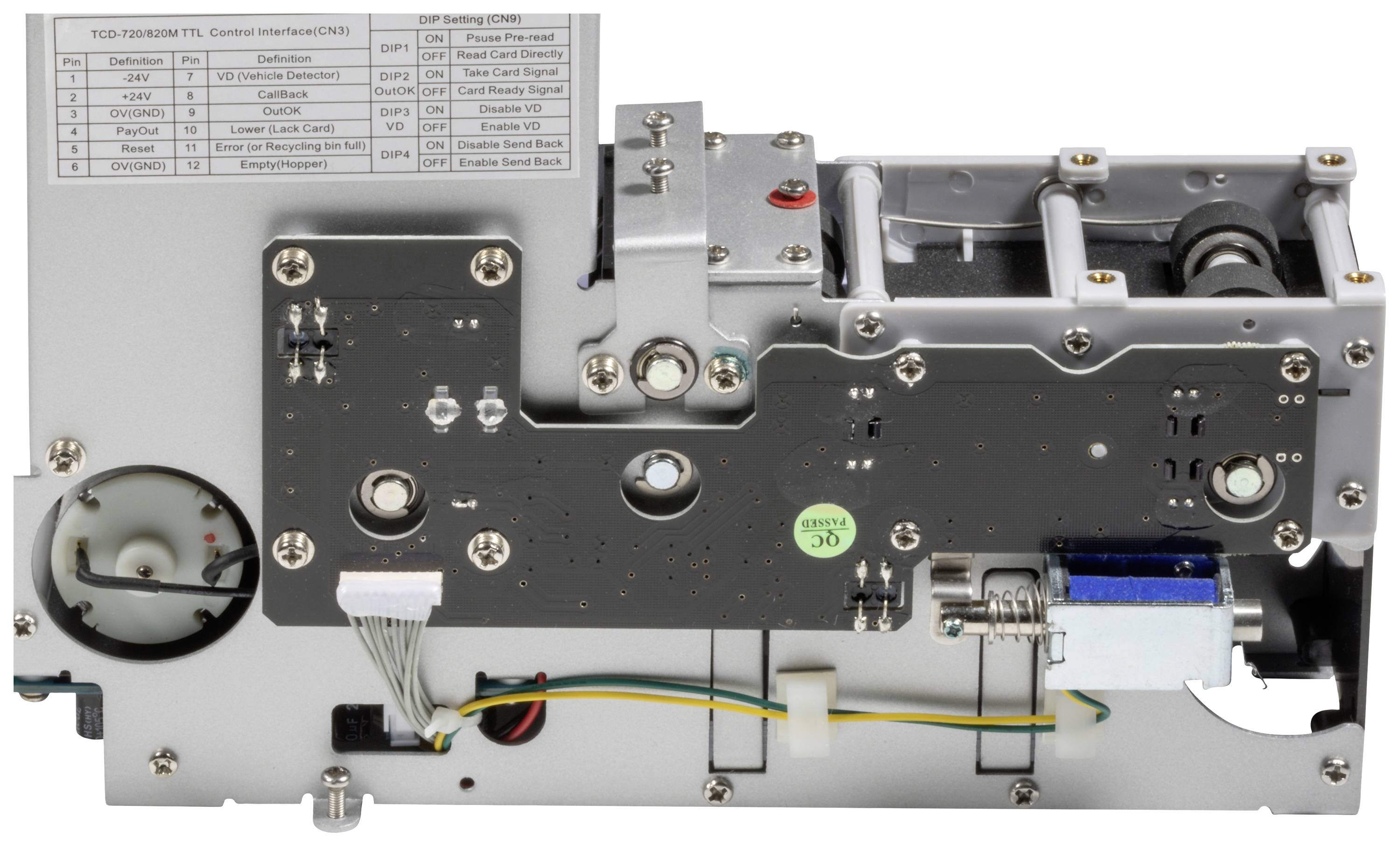 A grey mechanical device with multiple screws, gears, and cable connections; a table with labels located in the top left corner.