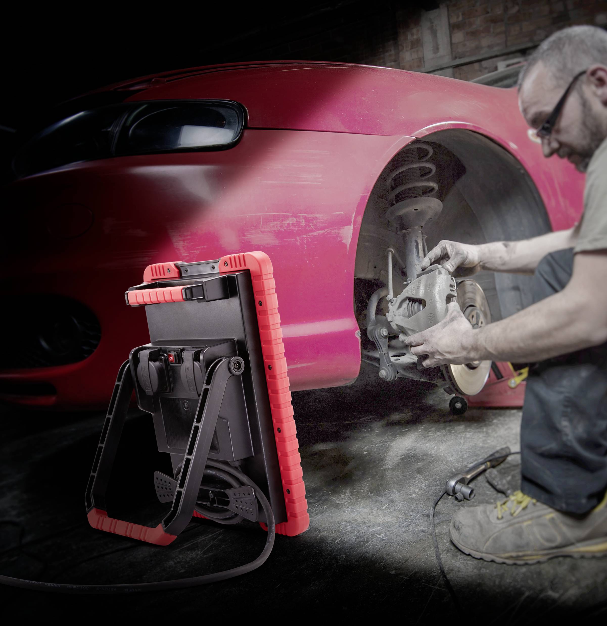 A mechanic is repairing the brakes of a red car. In front of him stands a bright work lamp illuminating the area.